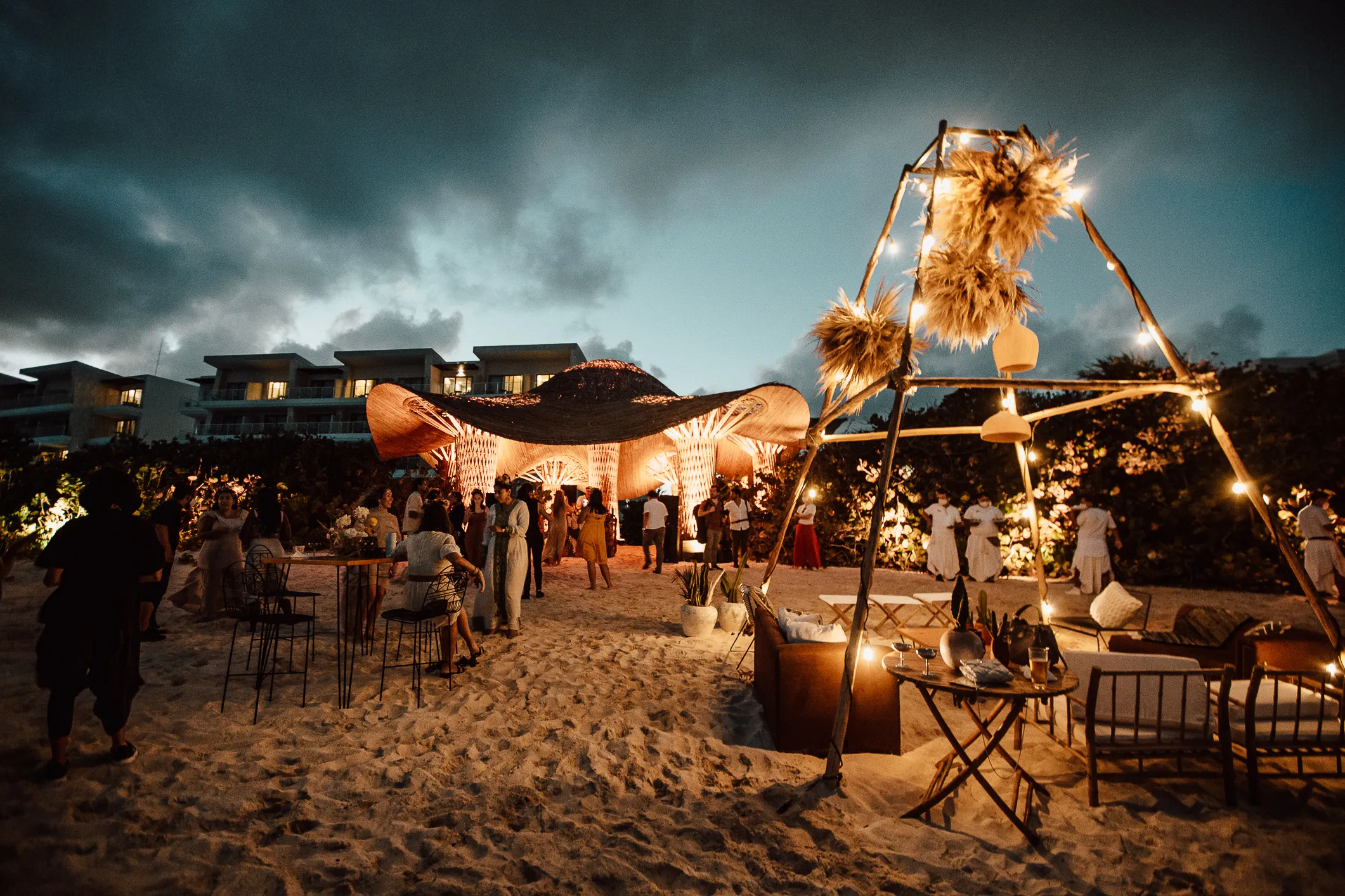 Evening beachfront gathering with guests around a lit sculptural pavilion and lounge seating.