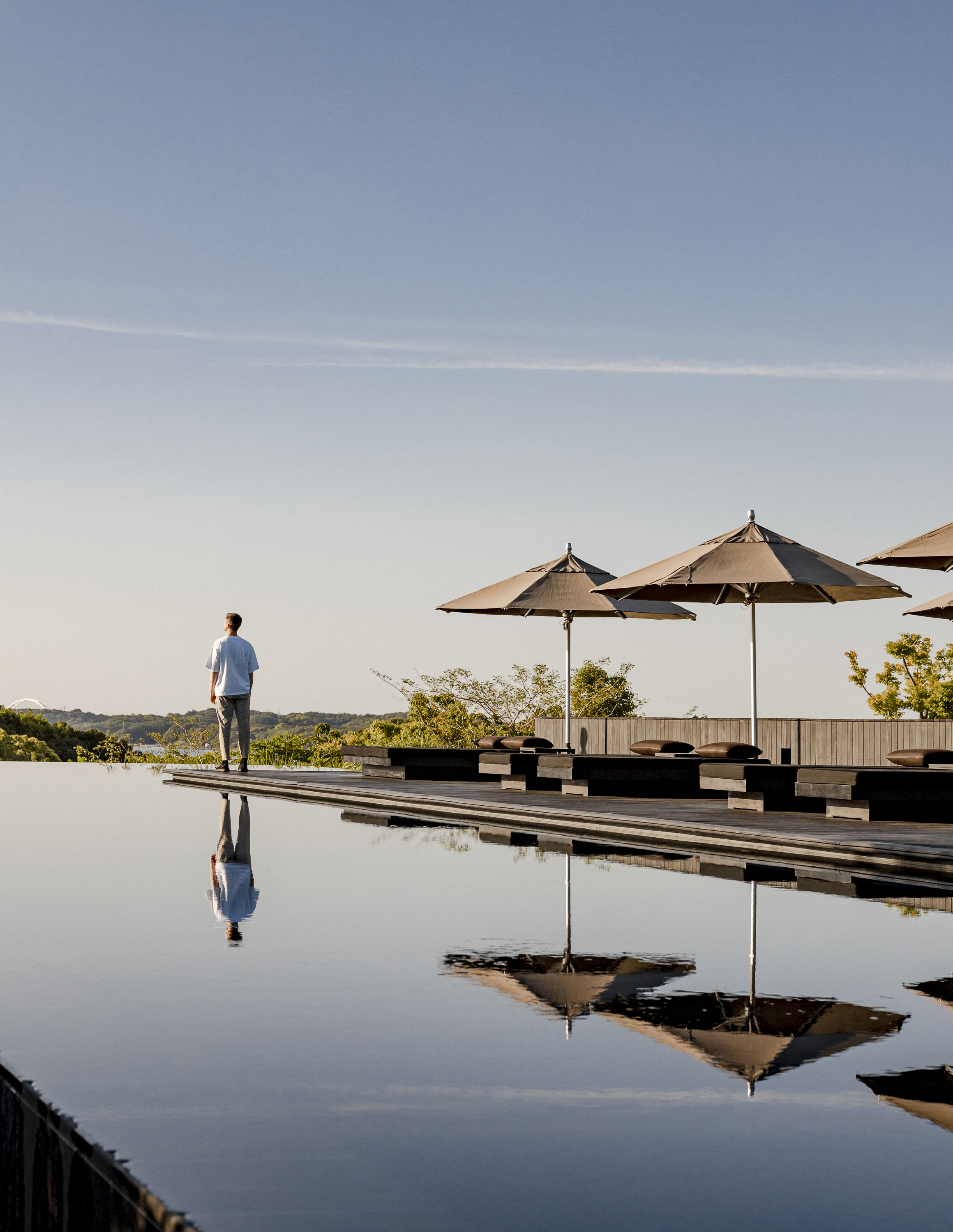 A man standing on the edge of an infinity pool looking at the view next to sun loungers reflected in the water