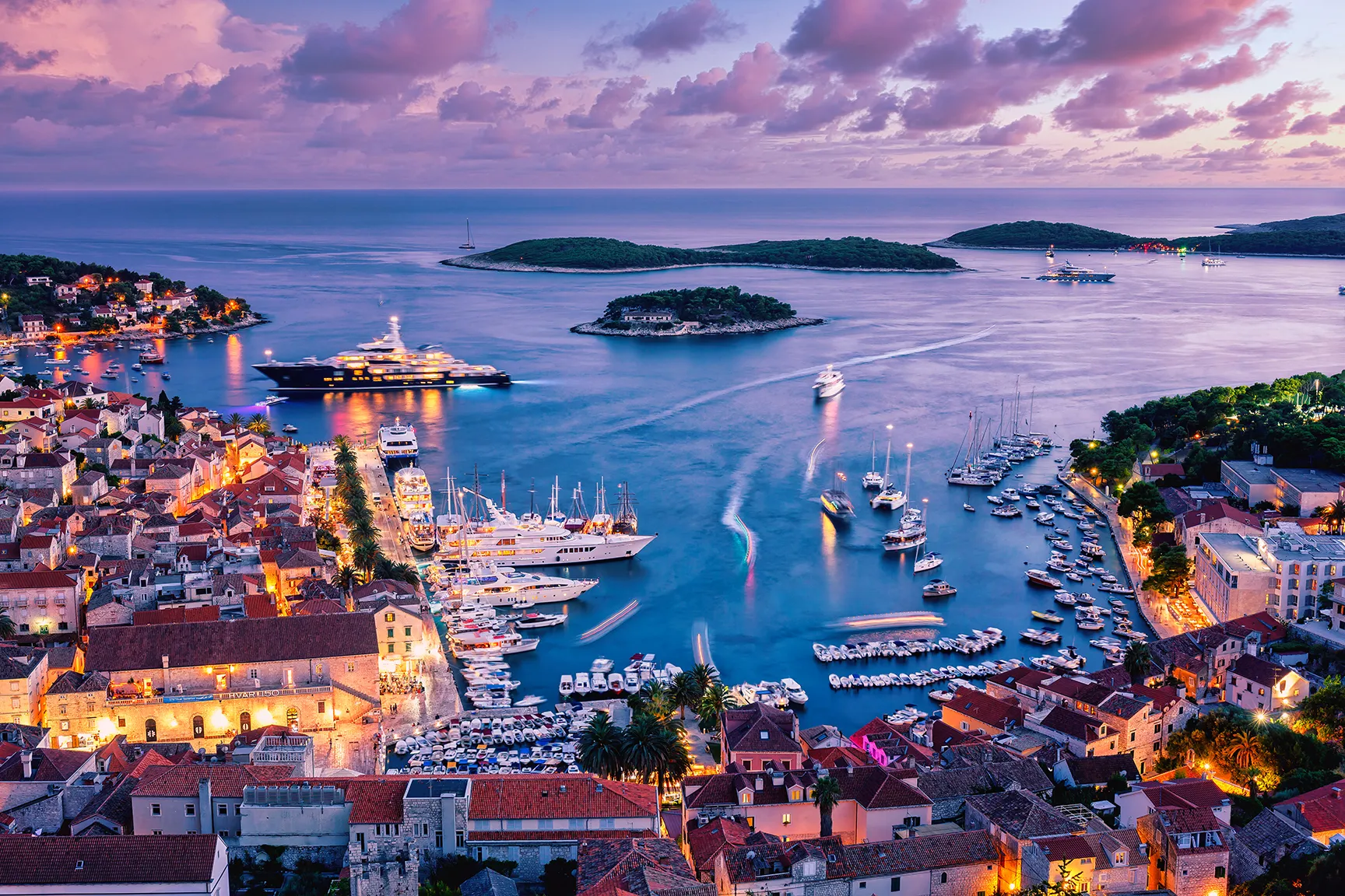 Twilight view of a coastal harbor with yachts, island-dotted waters, and illuminated waterfront buildings.