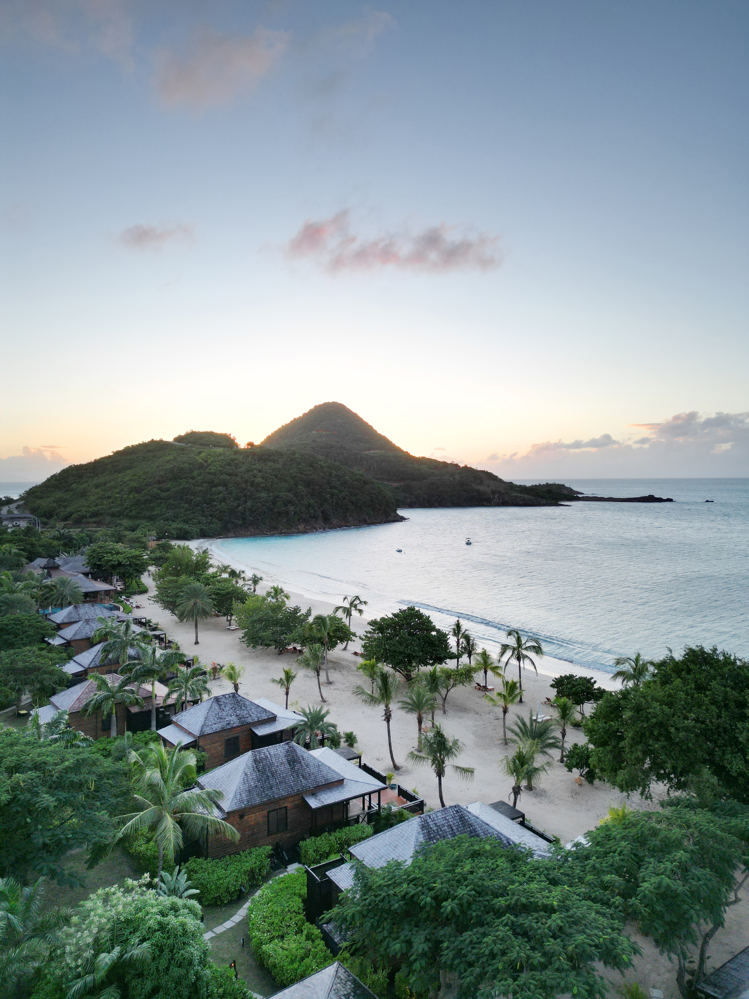 A birds eye view of Hermitage Bay in Antigua featuring villas by a beach studded with palms and mountains in the background