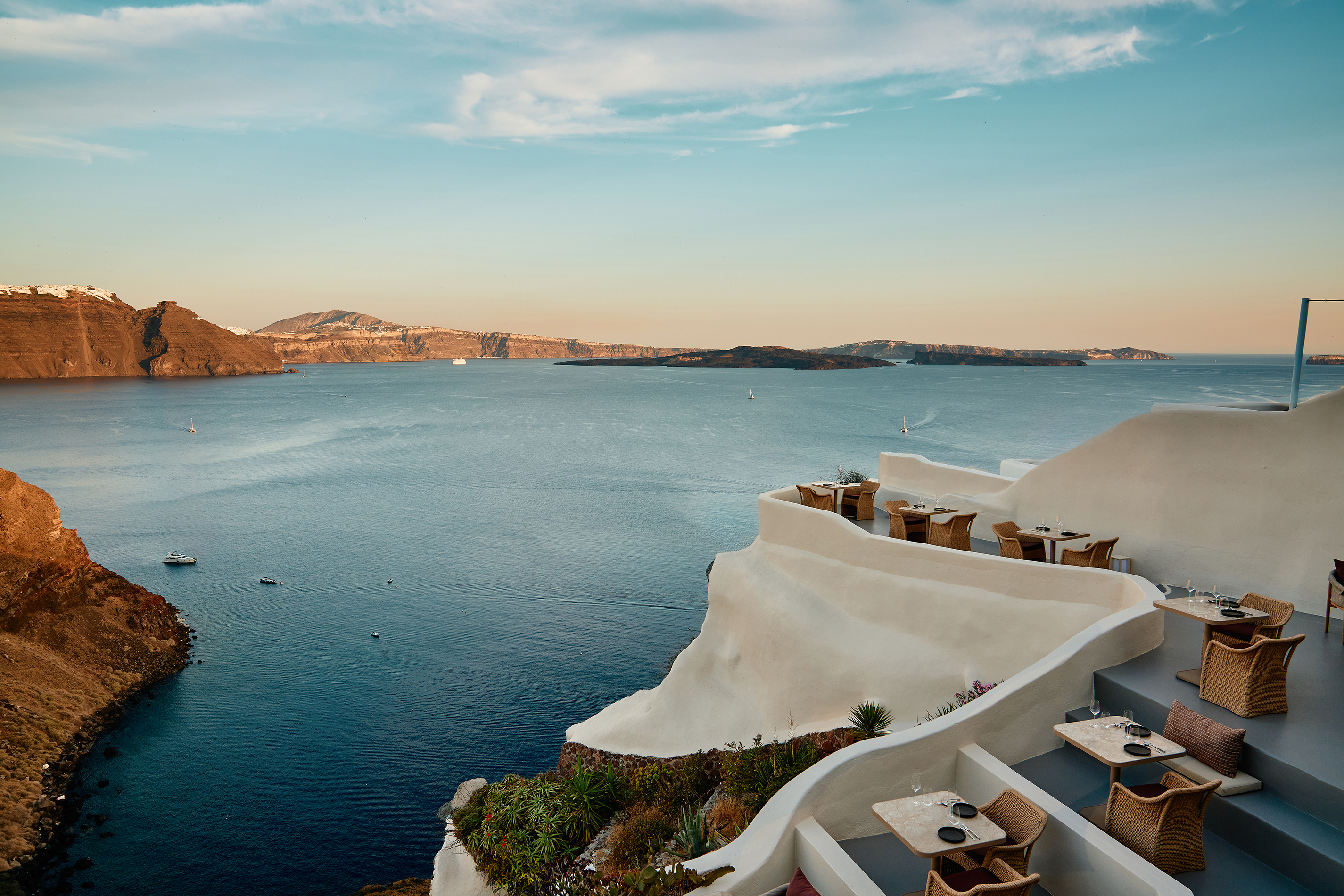 A white terrace with tables on the edge of a cliff facing a vast landscape of ocean and mountains