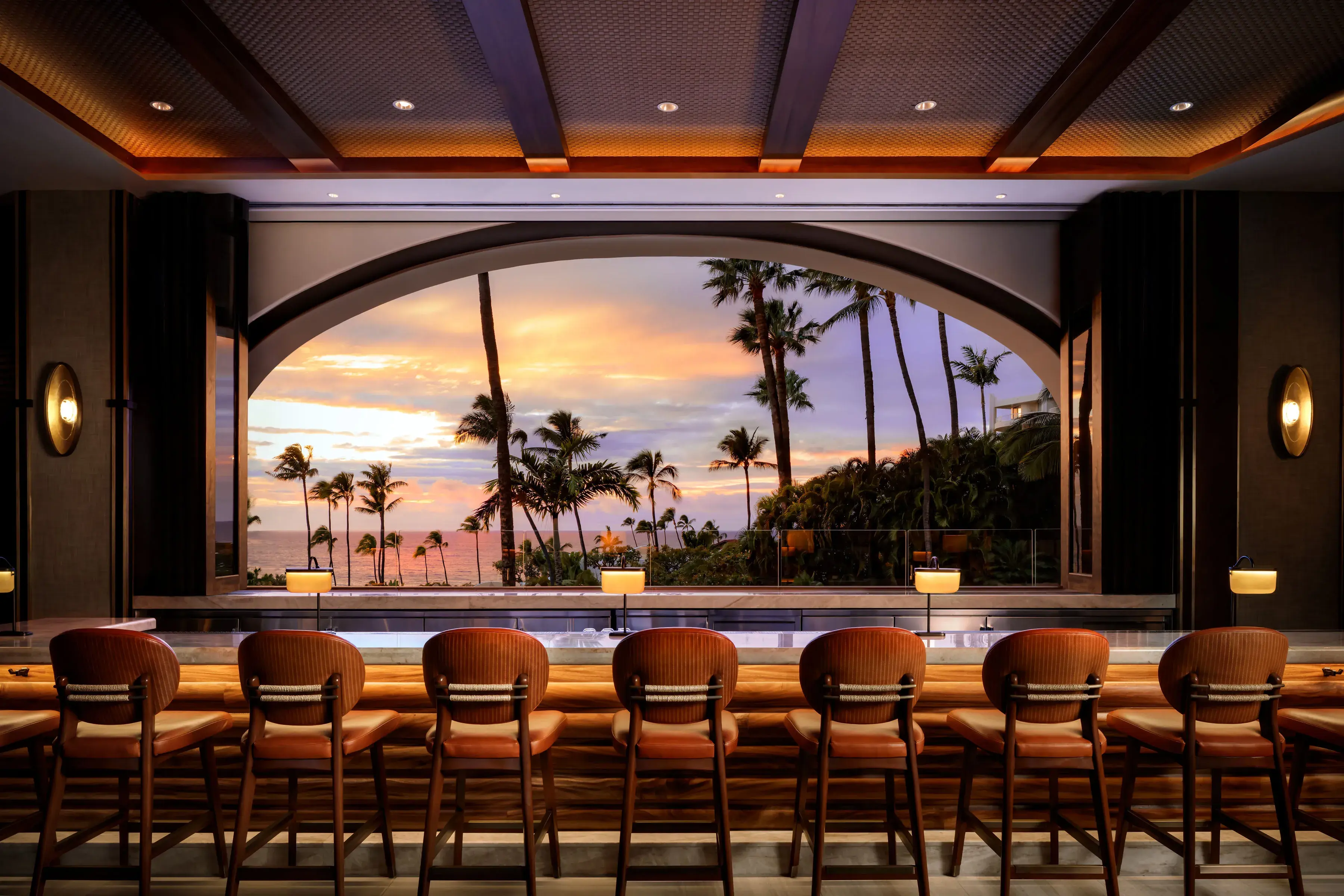 The moody bar area of PILINA Restaurant at  Fairmont Kea Lani with several bar stools lined facing a large window overlooking the sunset