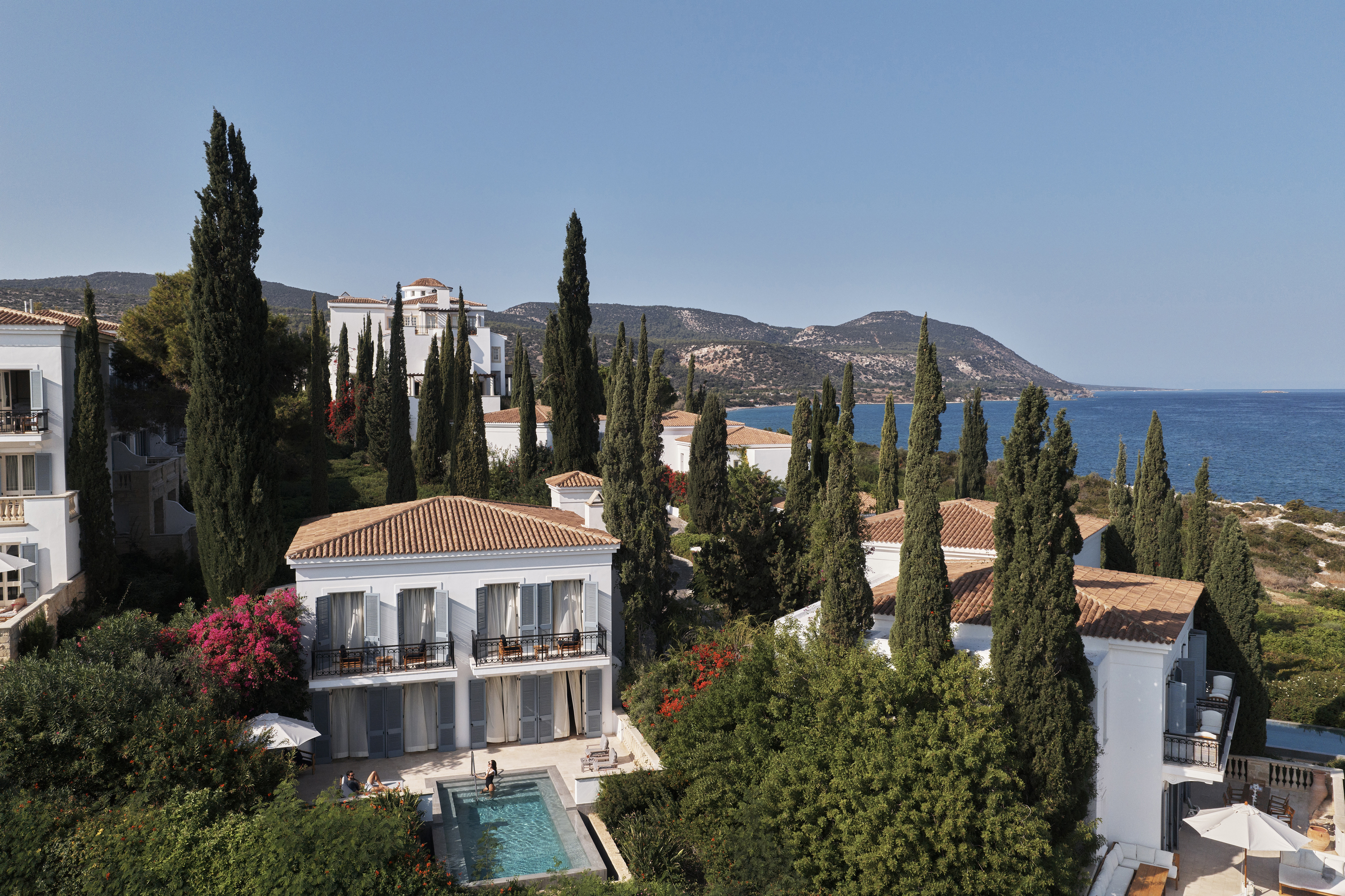 Aerial view of Alexandros Residence set among green trees and bushes with other villas surrounding it and hills behind