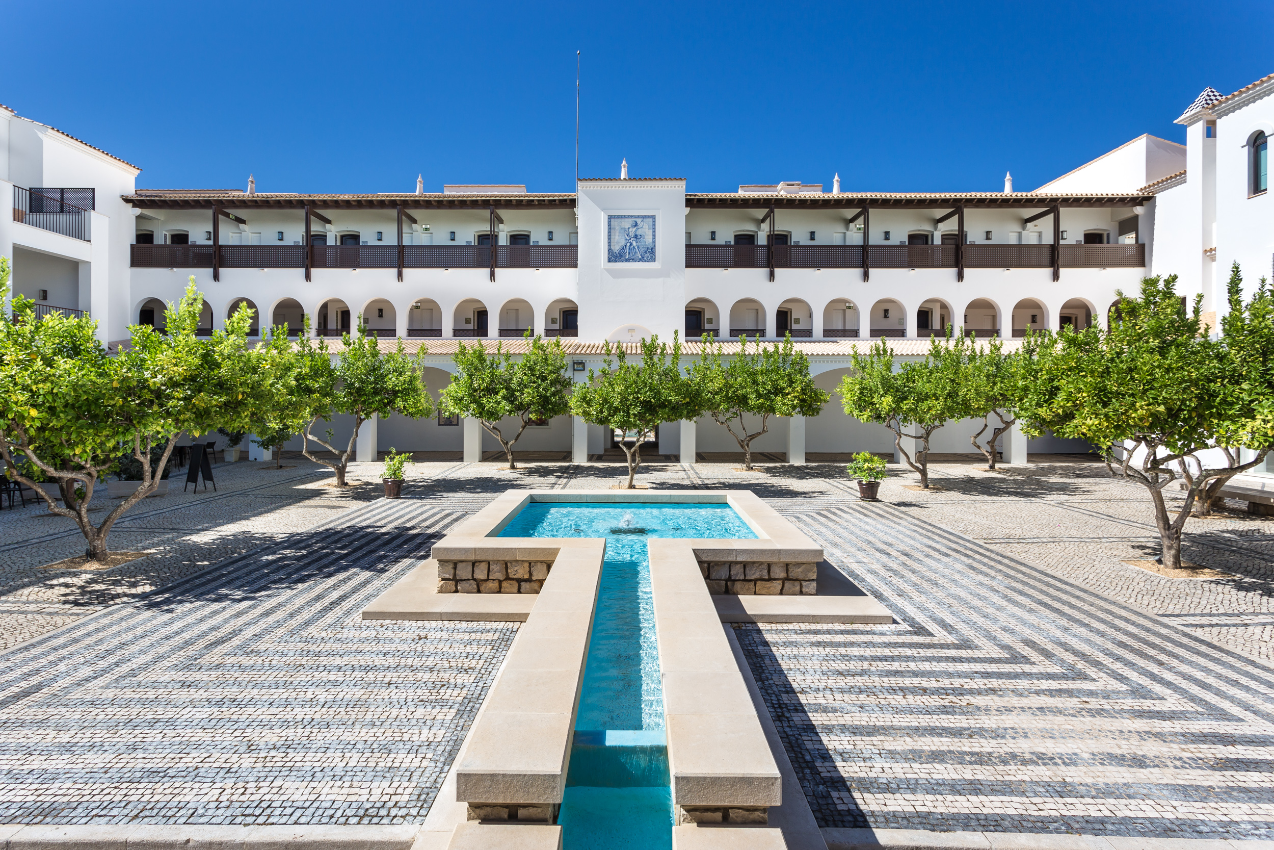 A courtyard with a central water feature, surrounded by trees and a building featuring arches and balconies under a clear blue sky