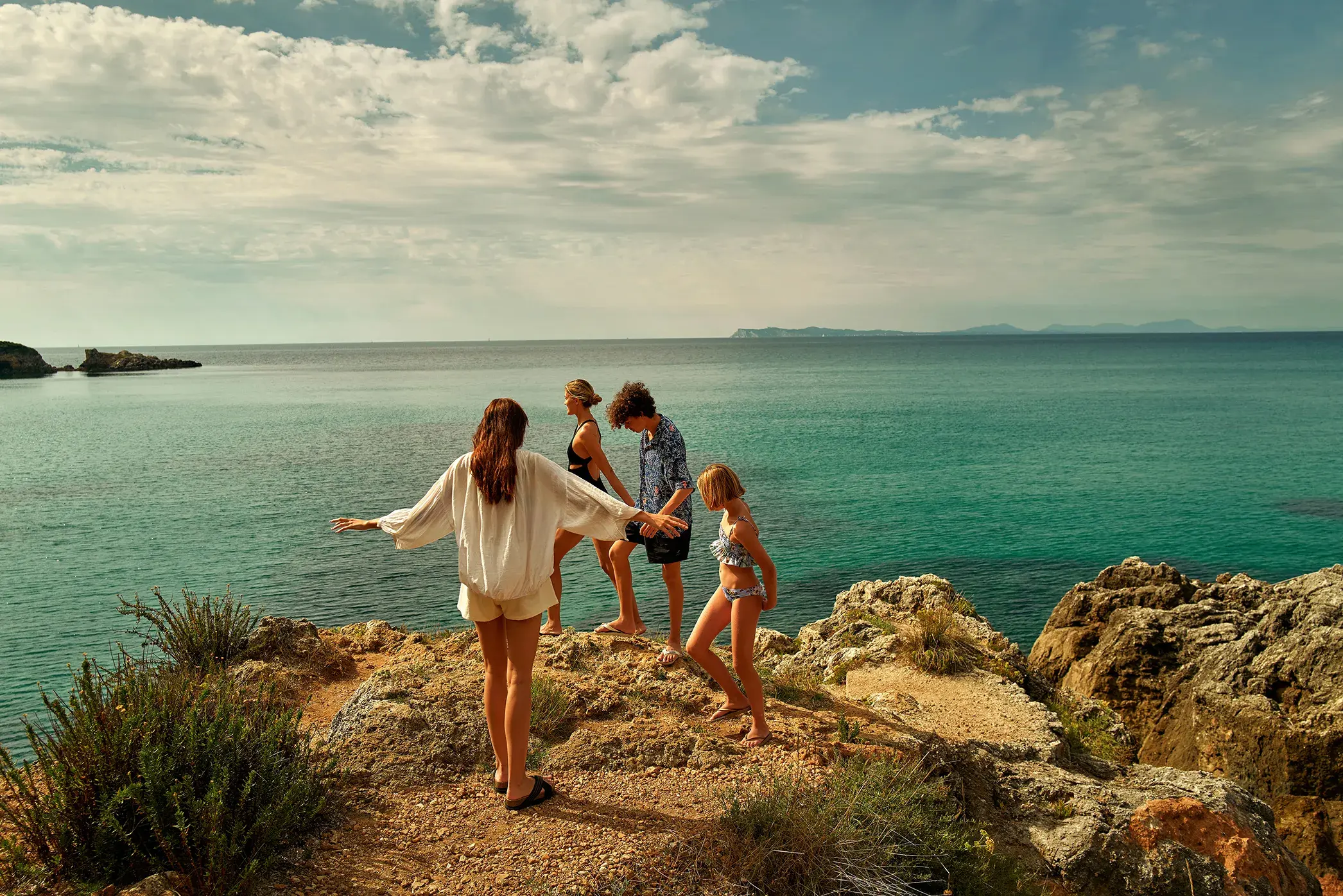 Group of people exploring rocky coastline with clear turquoise sea and distant islands under a partly cloudy sky.