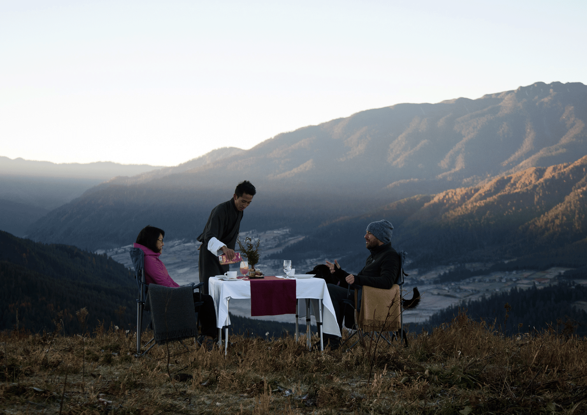 Two guests enjoying an outdoor dining experience on a mountain ridge with sweeping valley views.