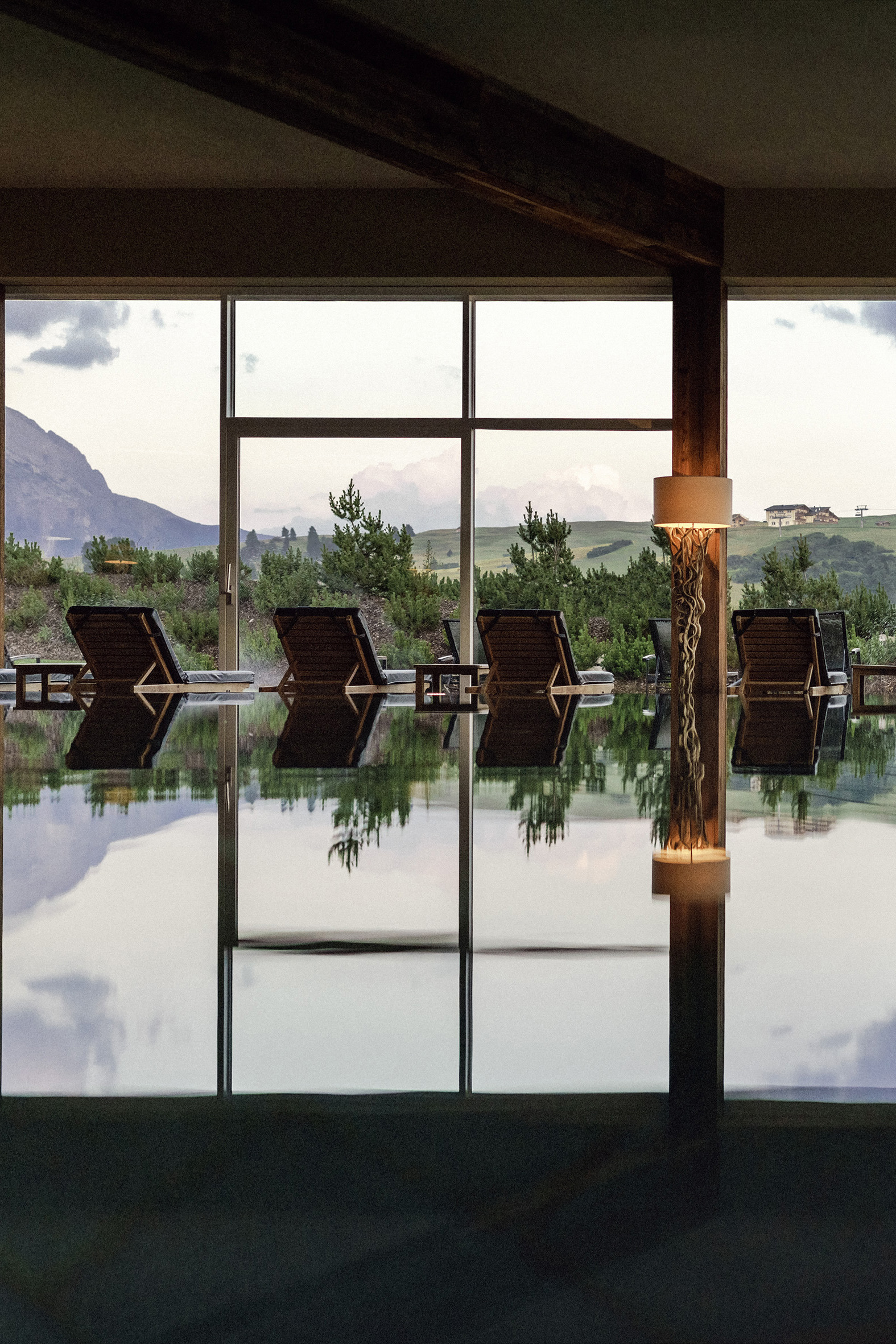 An indoor pool area with a row of lounge chairs facing large windows that offer a view of a serene landscape with mountains in the distance.