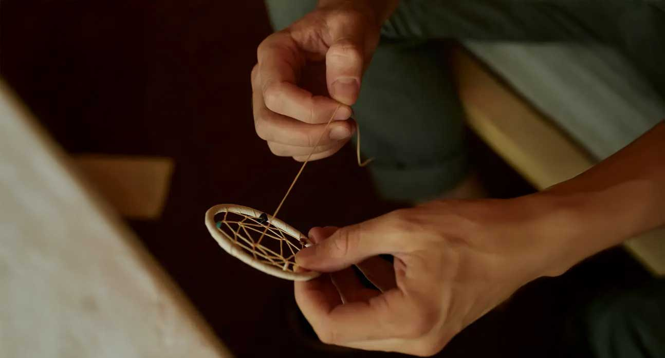 Close‑up of hands weaving a small dreamcatcher with fine thread in warm indoor light.