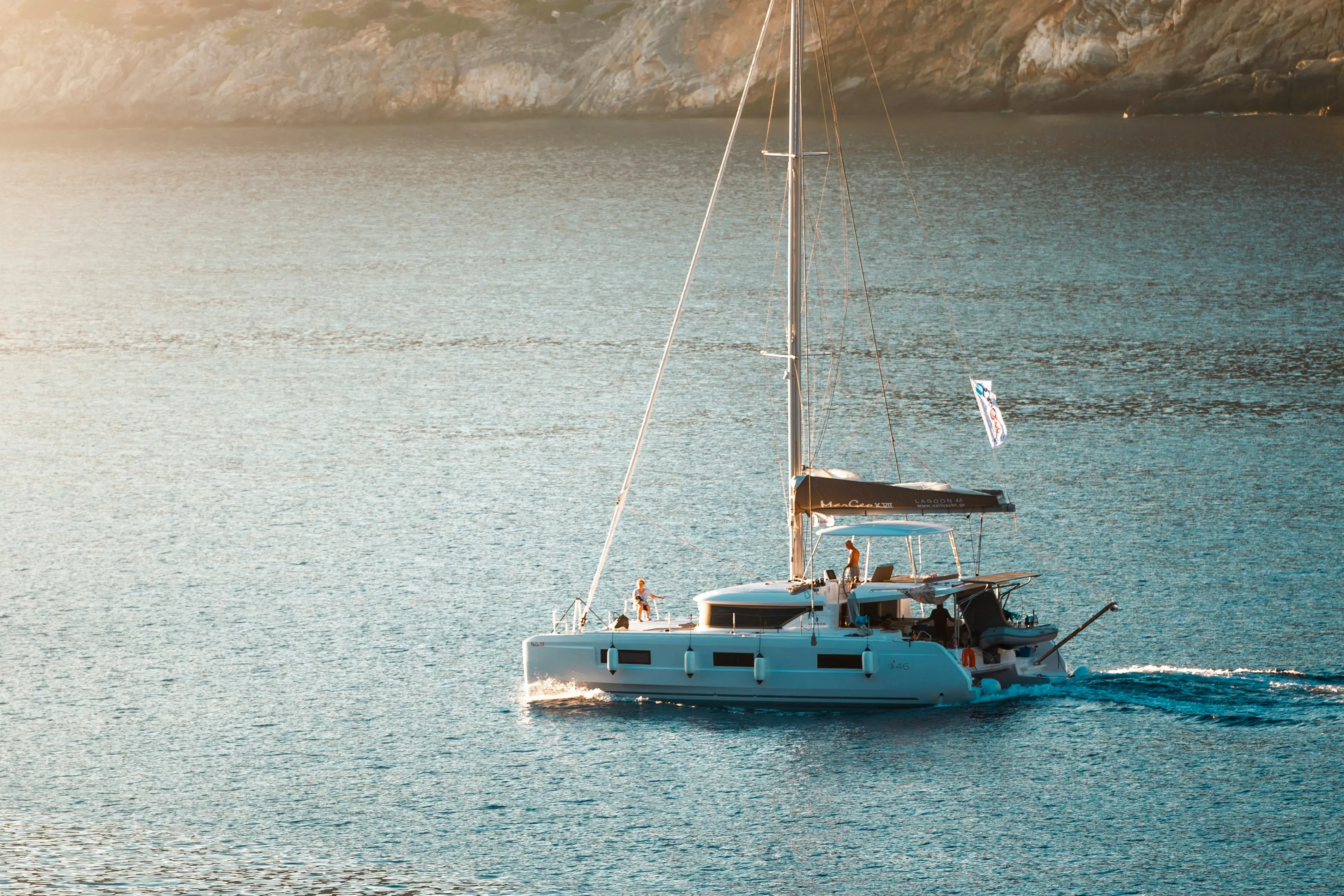 Modern white catamaran sailing on calm blue water near a rocky coastline at sunset.