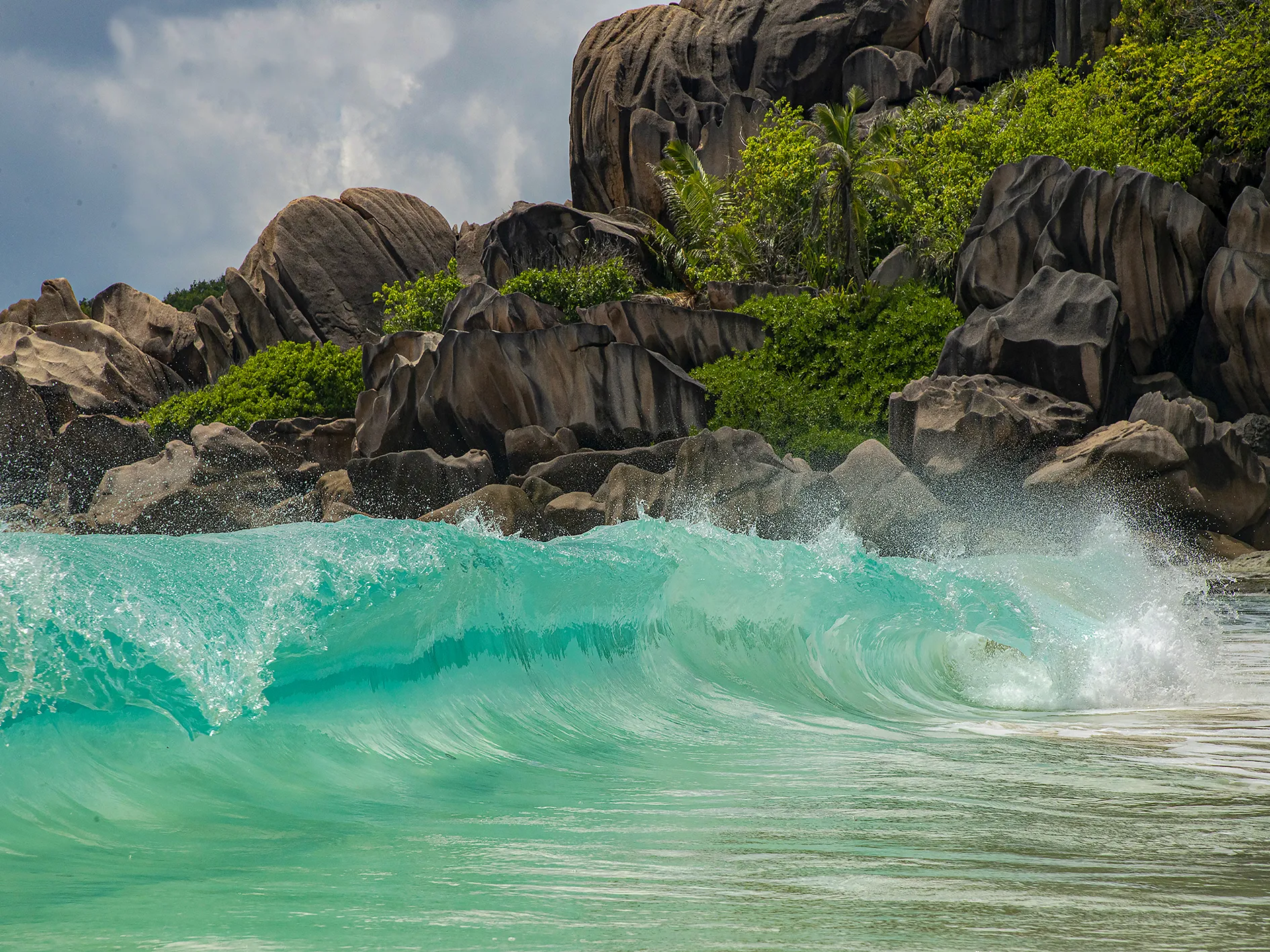Turquoise ocean wave crashing against dramatic granite rock formations and lush greenery on the tropical coastline of La Digue Island.