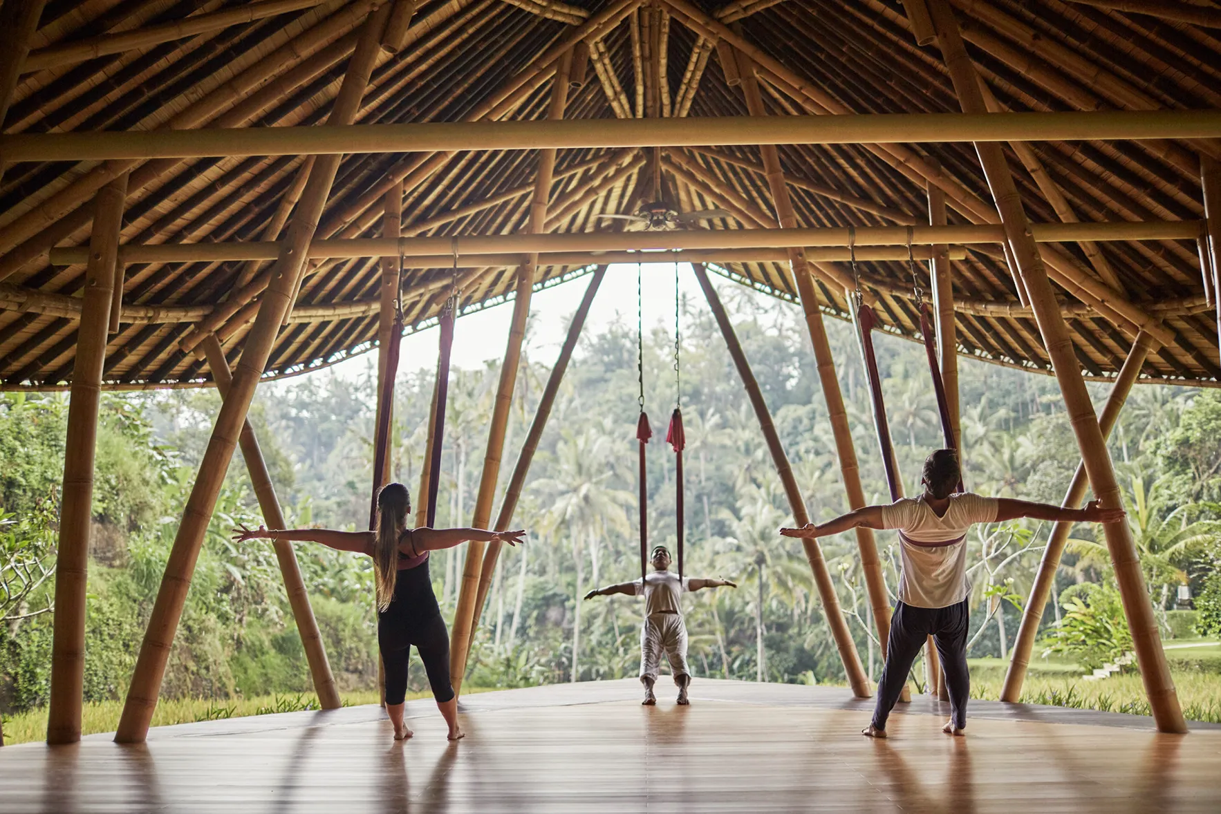 Three people practicing antigravity yoga, leaning into hammocks suspended from a bamboo roof of a pavilion at Four Seasons Resorts Bali at Sayan, Indonesia