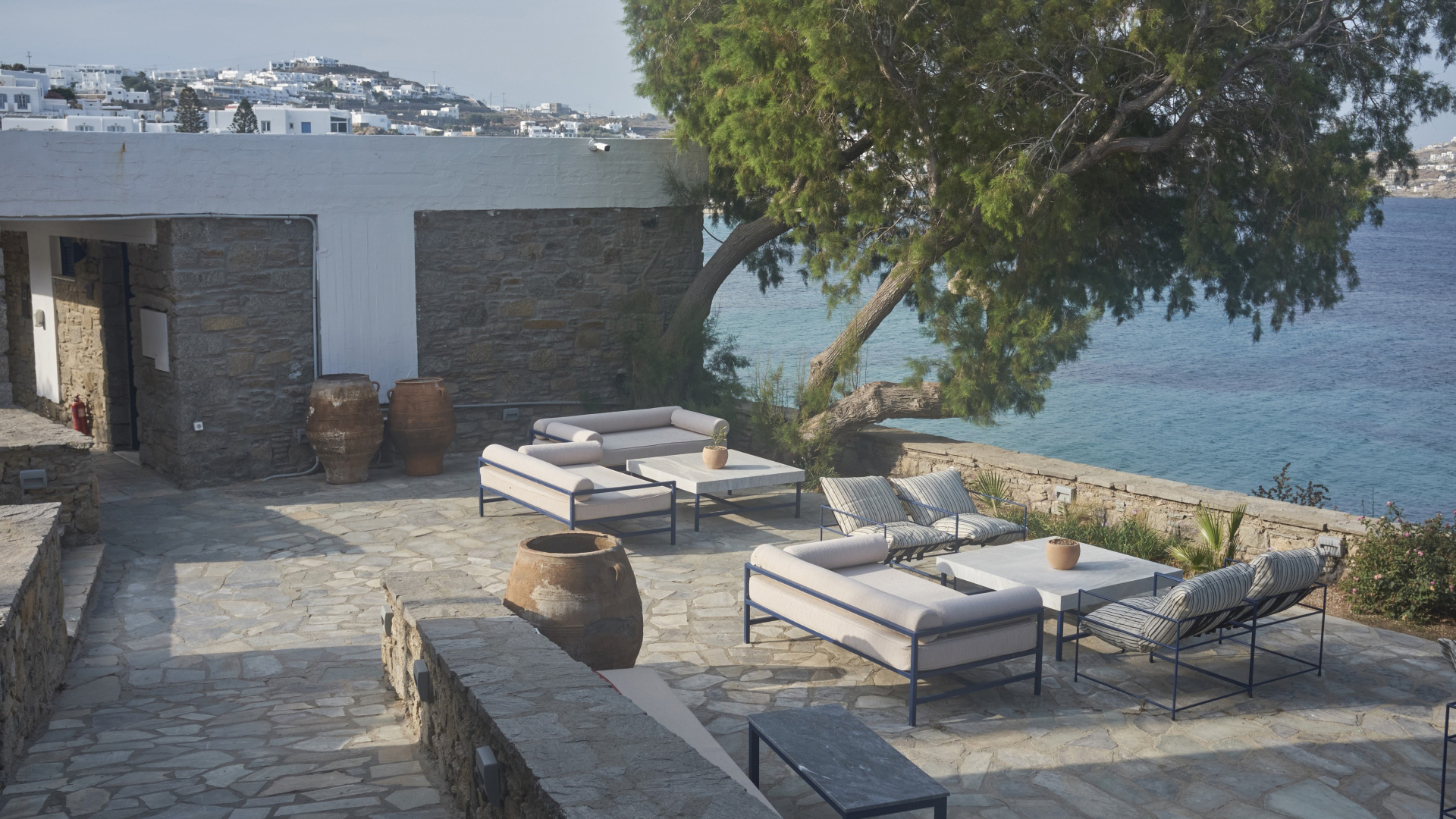 A stone patio with comfy furniture and decorative pots below a large tree beside the sea