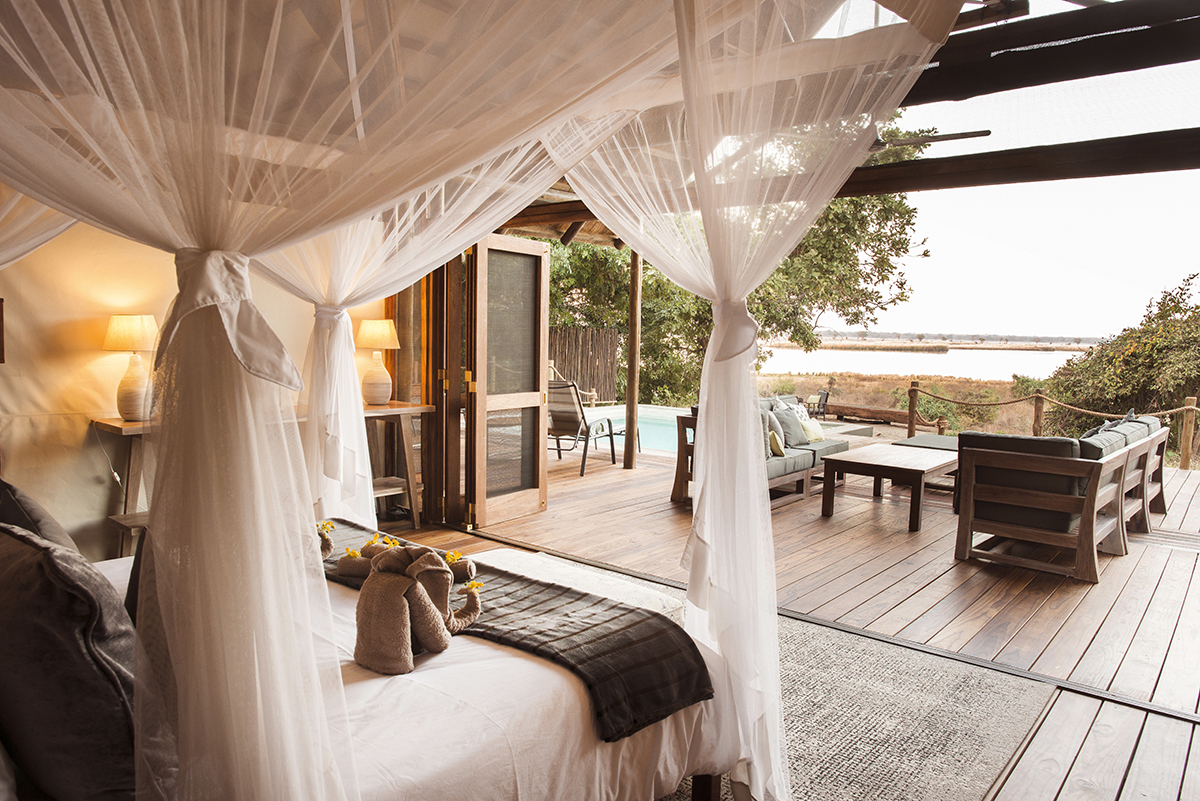 A Safari Suite bedroom at Chiawa Camp overlooking the Zambezi River featuring a four poster bed with white nets, wooden flooring and large doors pulled back leading onto a deck with outdoor seating