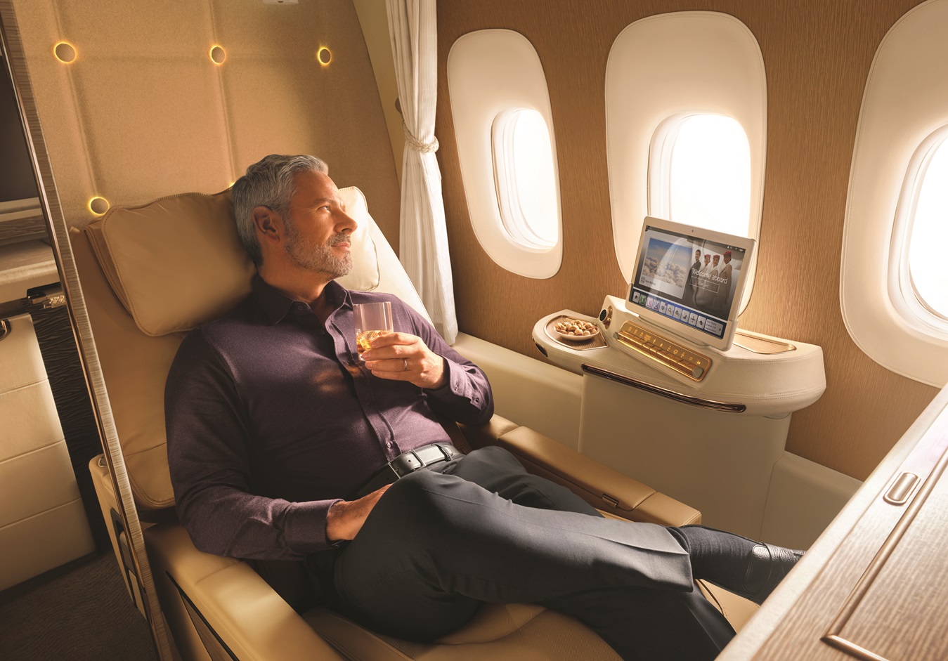 A person is reclining in a luxurious airplane seat, holding a drink and watching a screen.