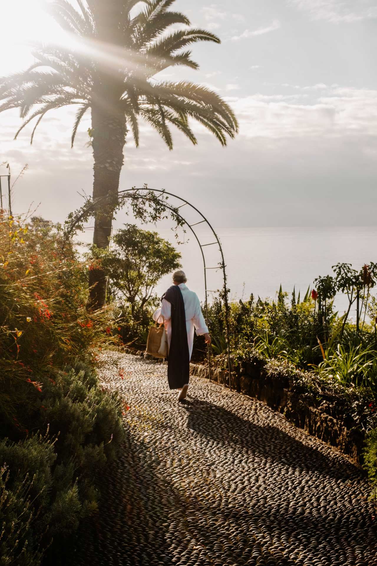 Guest walking along a sunlit garden path at a coastal resort, surrounded by lush greenery and palm trees overlooking the sea.