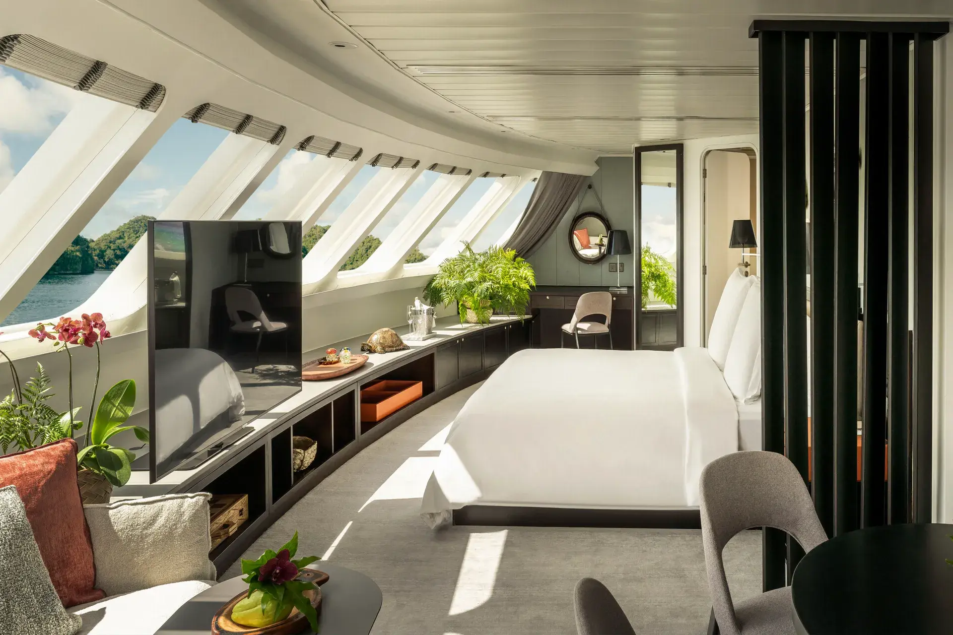 The bedroom of the Explorer Suite aboard Four Seasons Explorer Palau featuring a comfy bed, television and shelving unit with decorative ornaments in front of large curved windows