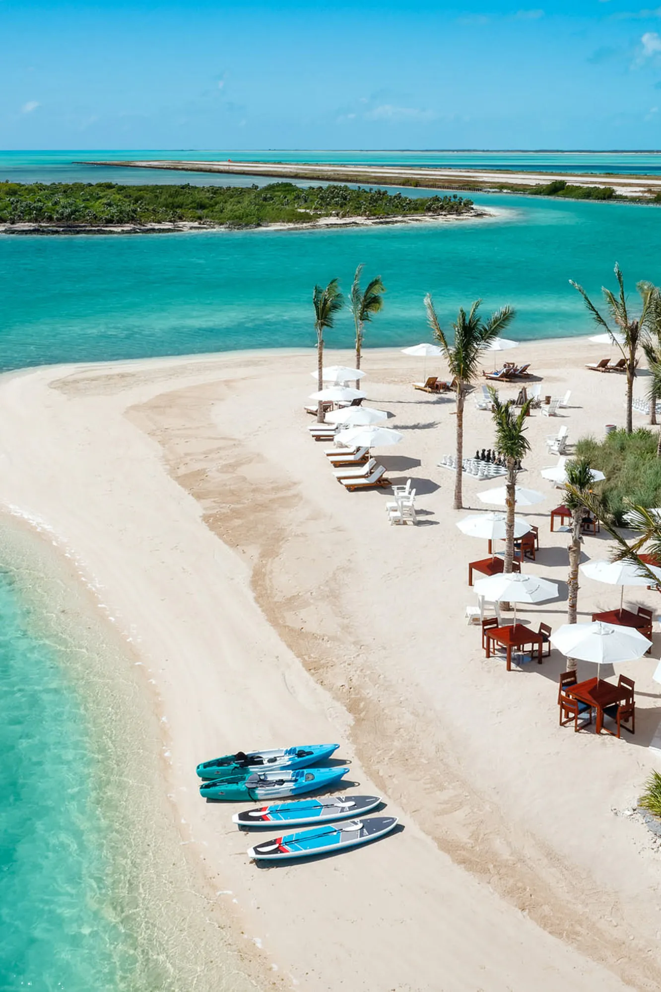 Aerial view of the Club House at Ambergris Cay in Turks & Caicos featuring water sports equipment at the water's edge and beach dining tables scattered between palms