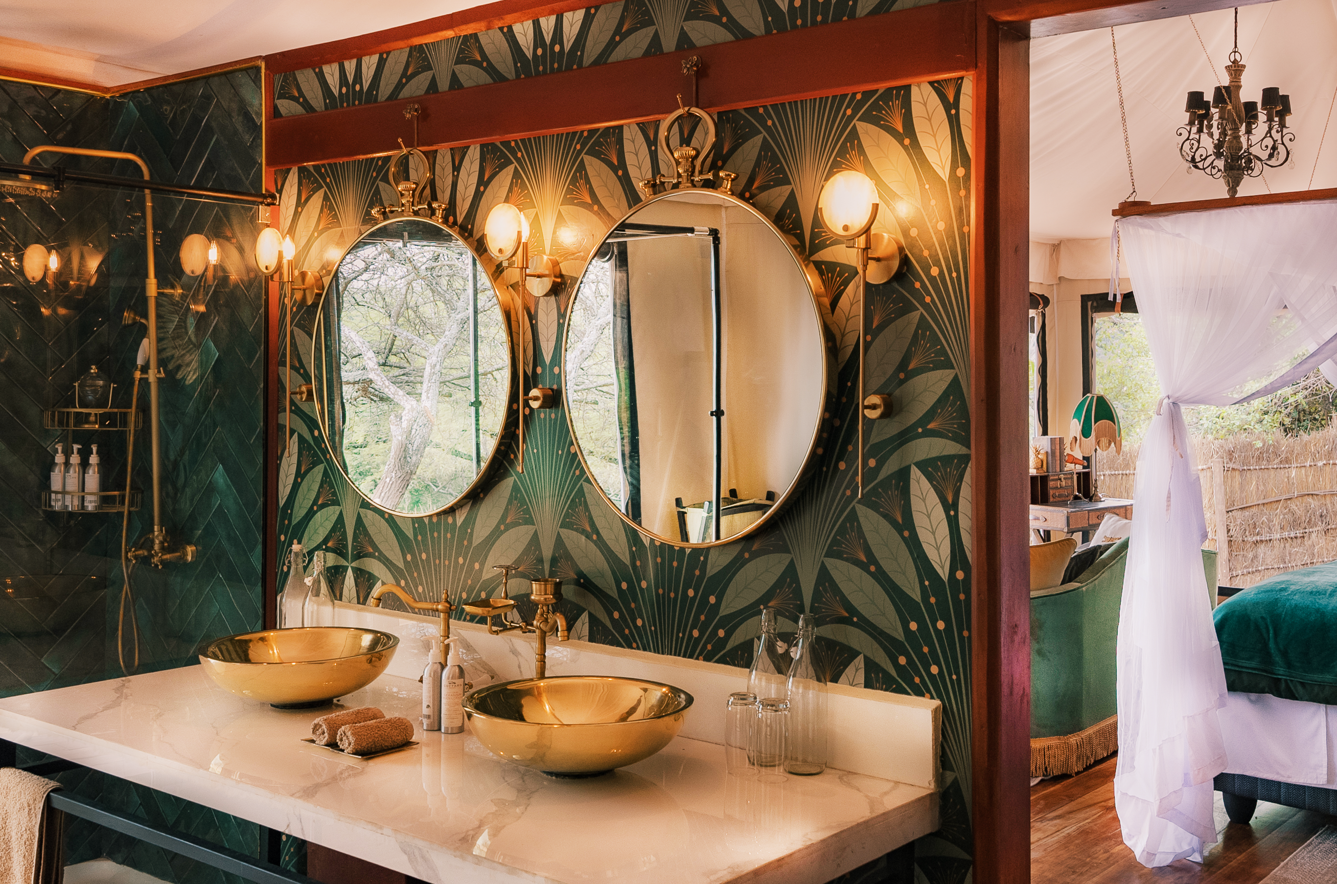 A luxurious bathroom at Cottars 1920s Camp featuring two gold sinks, round mirrors and art deco wallpaper with a glimpse into a bedroom with a canopy bed