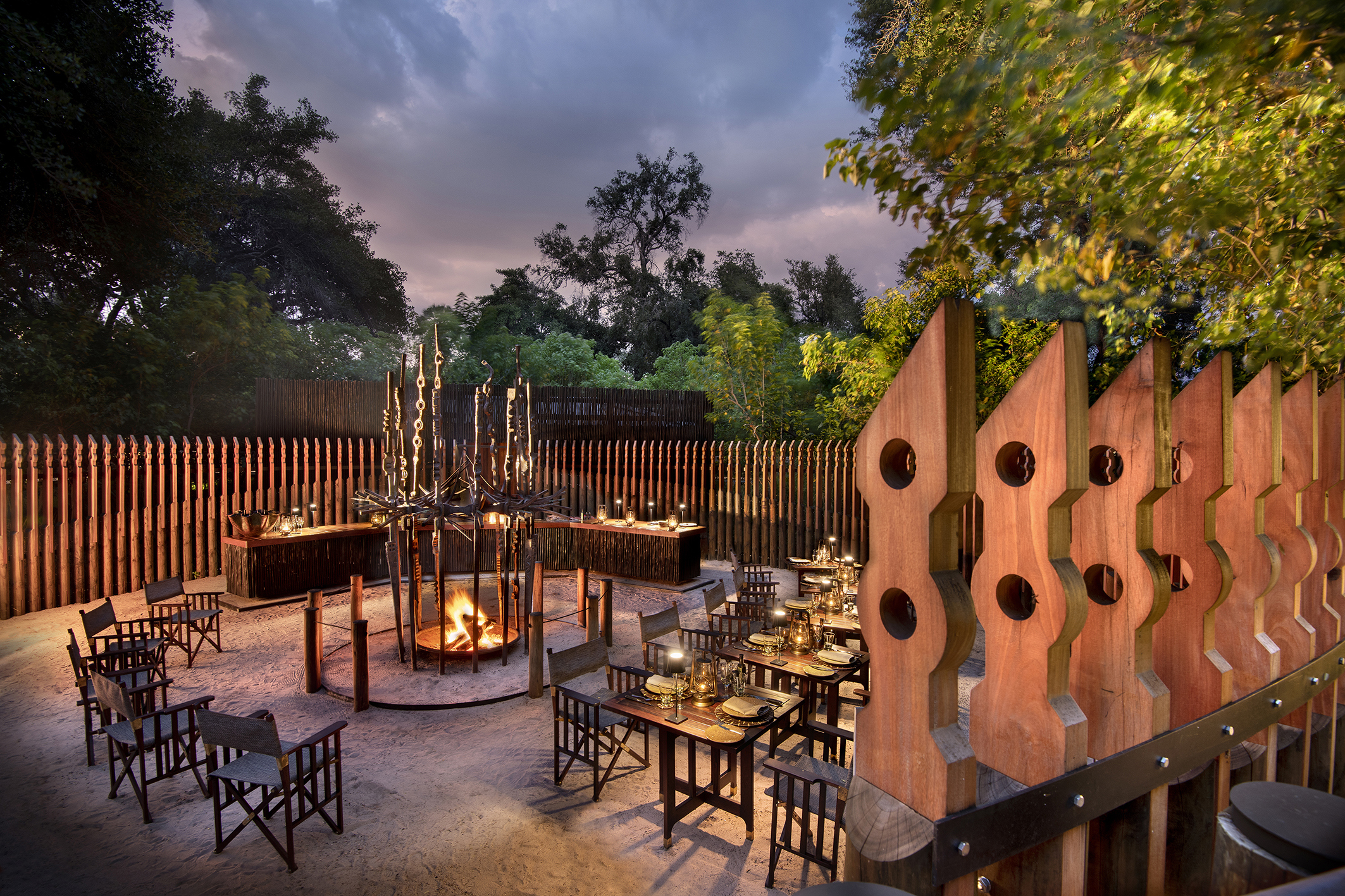 Five tables and a group of chairs set up at dusk for a Boma dinner with a lit fire pit in the centre and wooden sculptures 