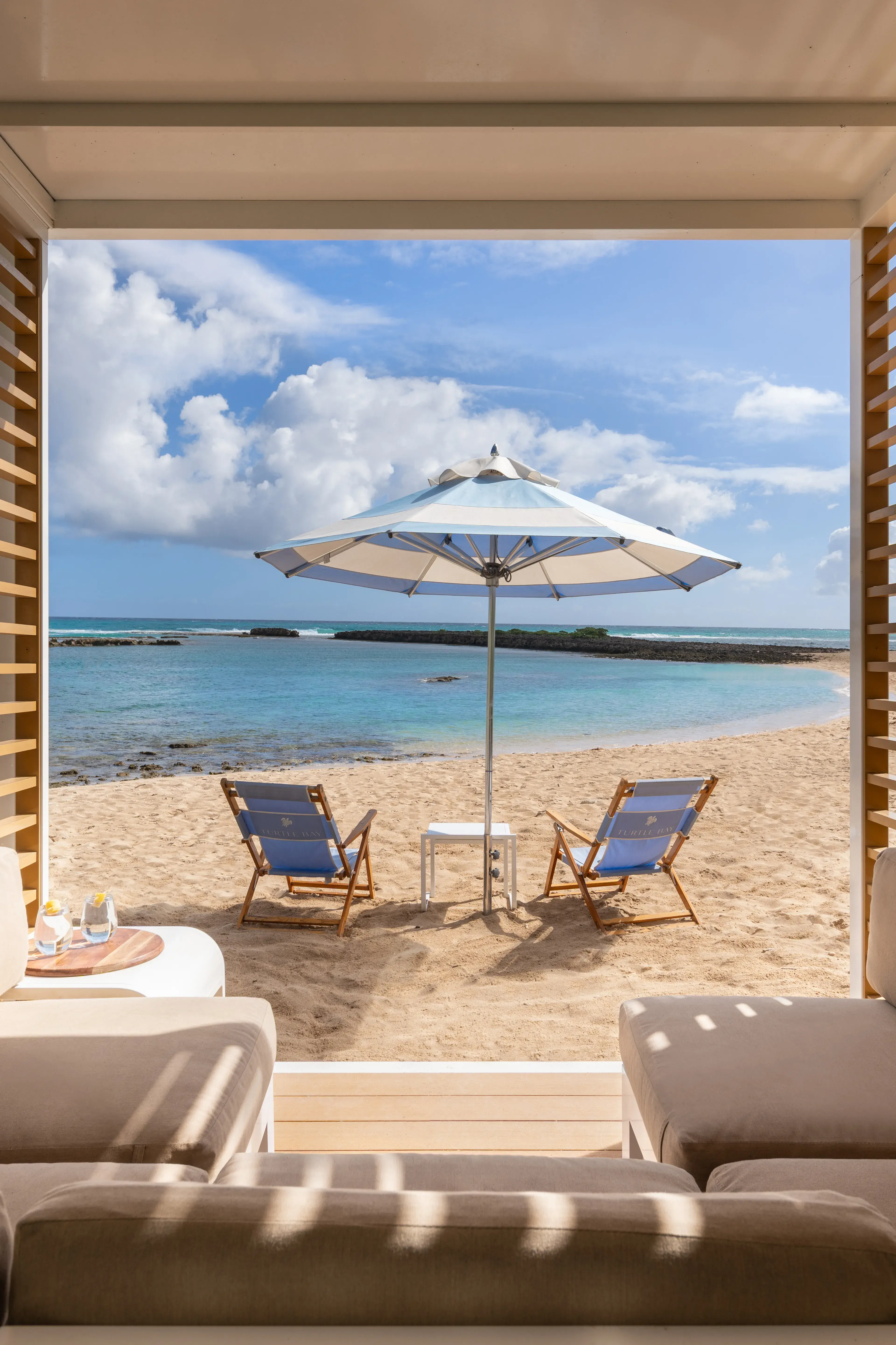 A cabana at the Beach Club at Ritz-Carlton Turtle Bay with comfy seating in the foreground looking out to sun loungers under and umbrella on the beach with the ocean in front