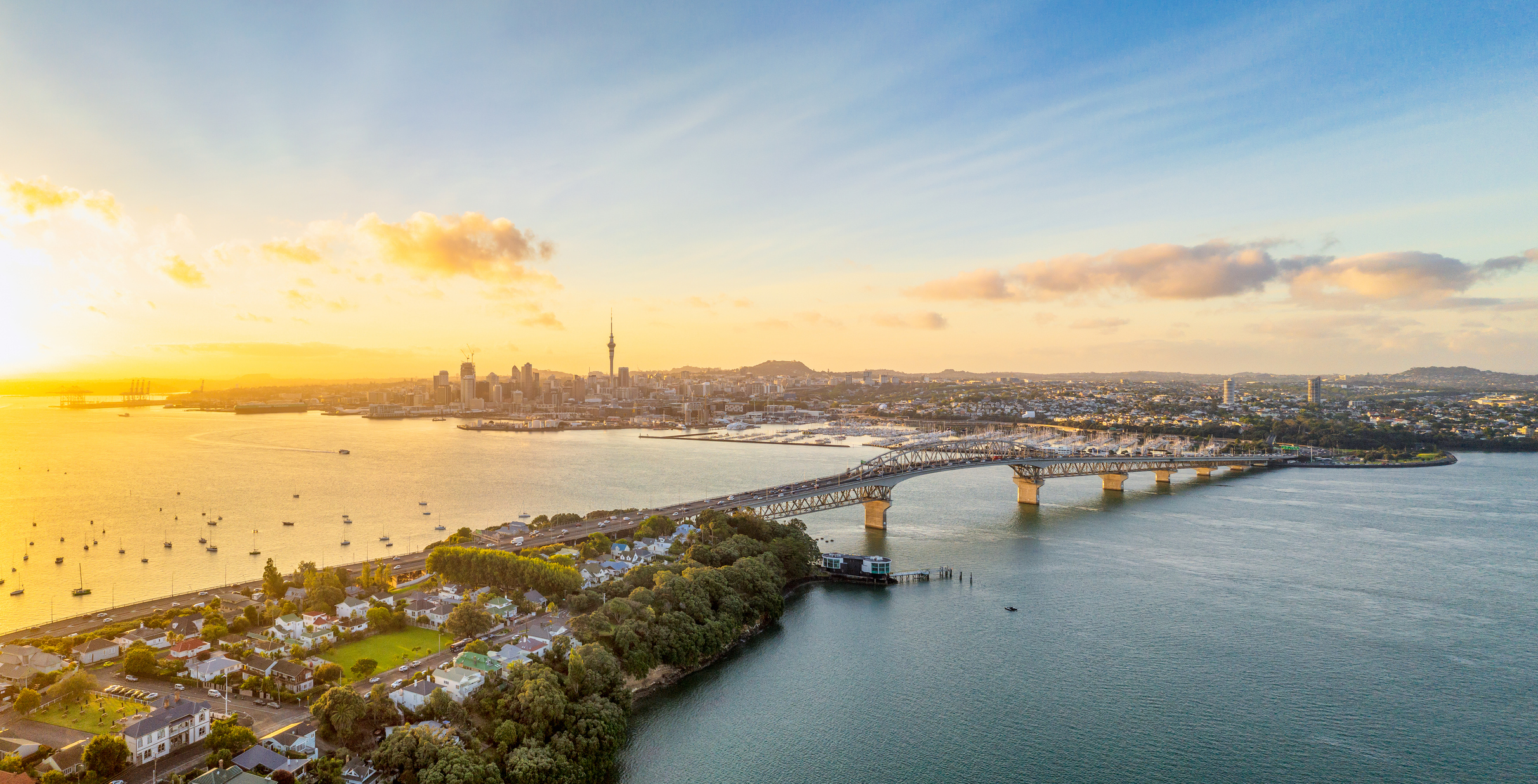 Aerial view of a city skyline at sunset with a bridge over water and boats docked along the shore.