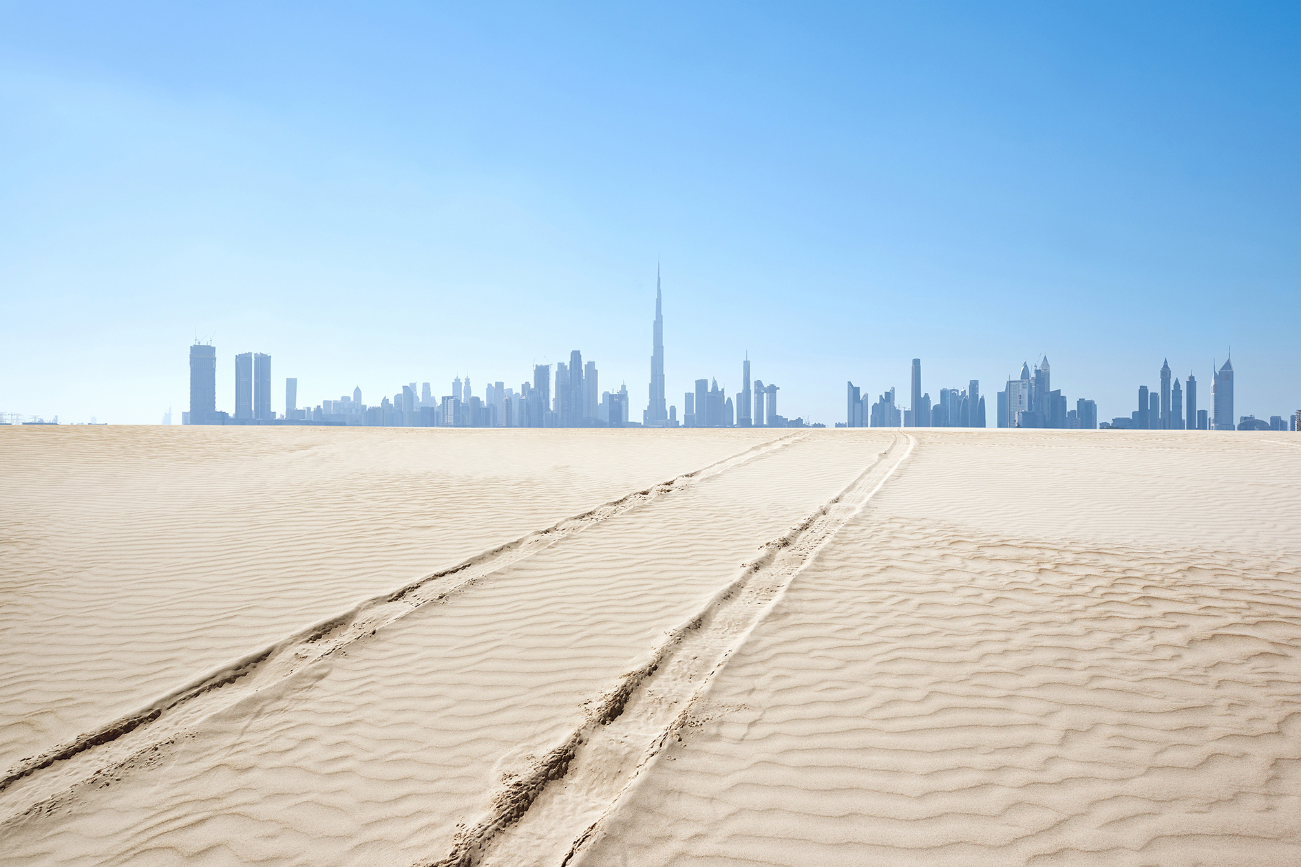 Tracks in white desert sand leading to Dubai skyline in background