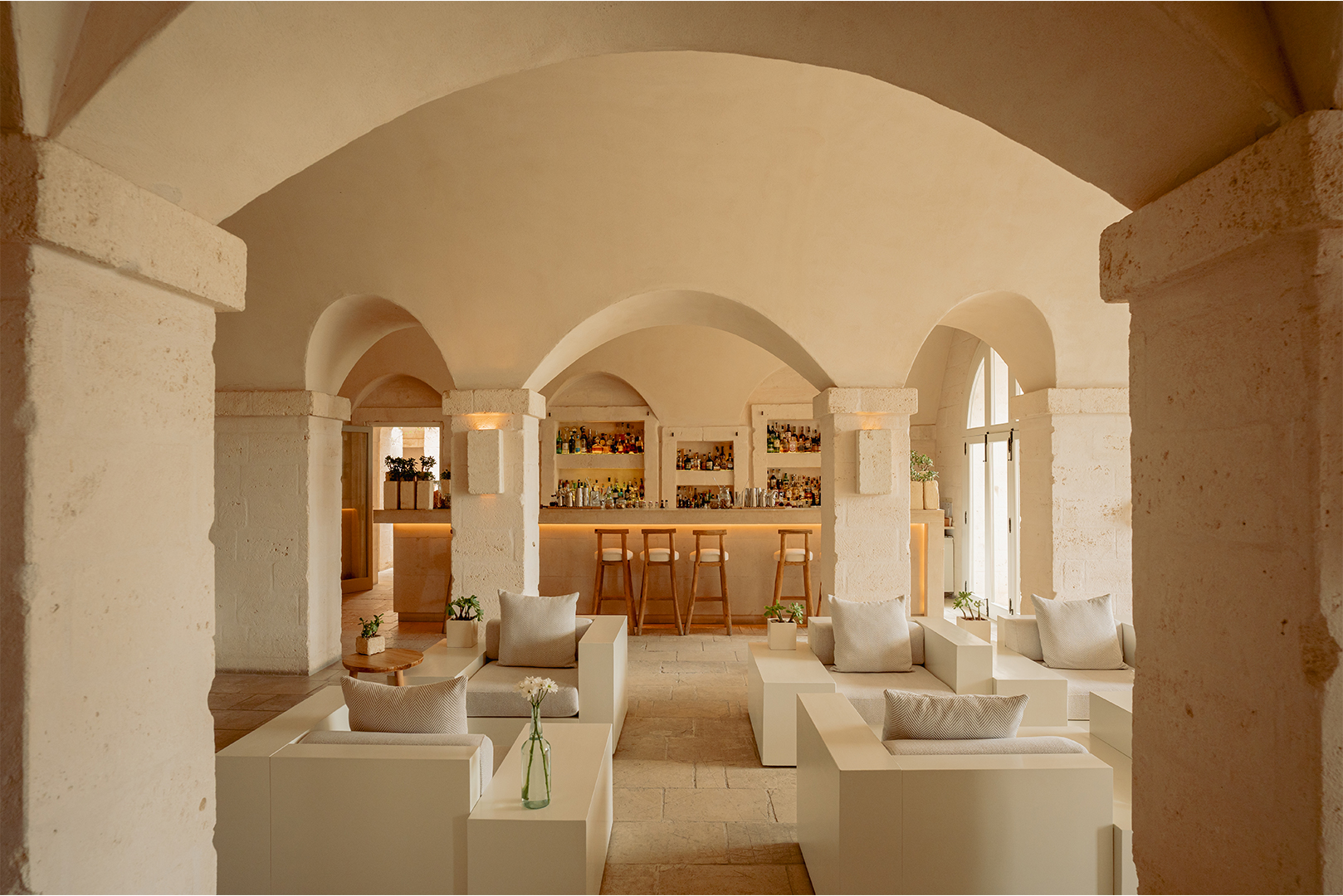 An elegant interior space with arched ceilings, white walls, and a series of tables with chairs and sofas arranged neatly, featuring a bar area with shelves of bottles in the background.