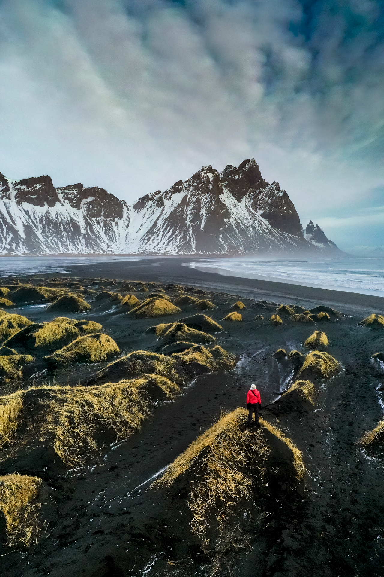A person standing on a black sand dune looking at Stokksnes bay with Vestrahorn mountain in the background.