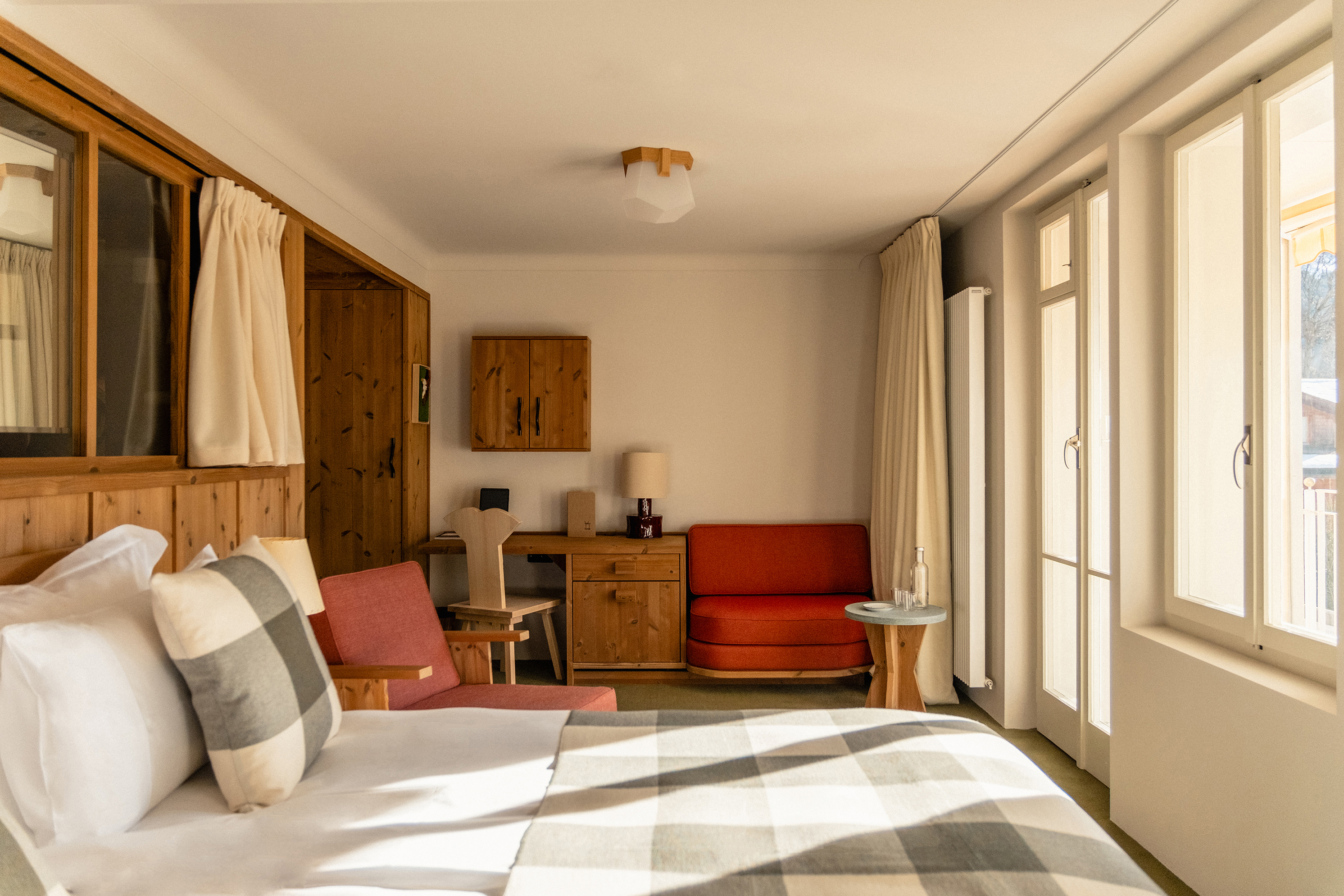 A superior room at Grand Hotel Belvedere featuring a bed with check details and a small lounge and desk area, with one wall of windows and doors and one of wooden panel details