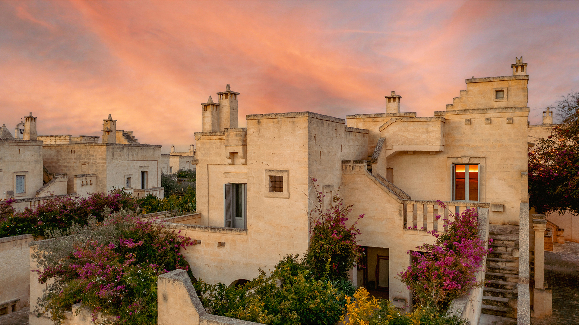 A photograph of ancient limestone buildings with staircases and blooming purple flowers under a sunset sky.