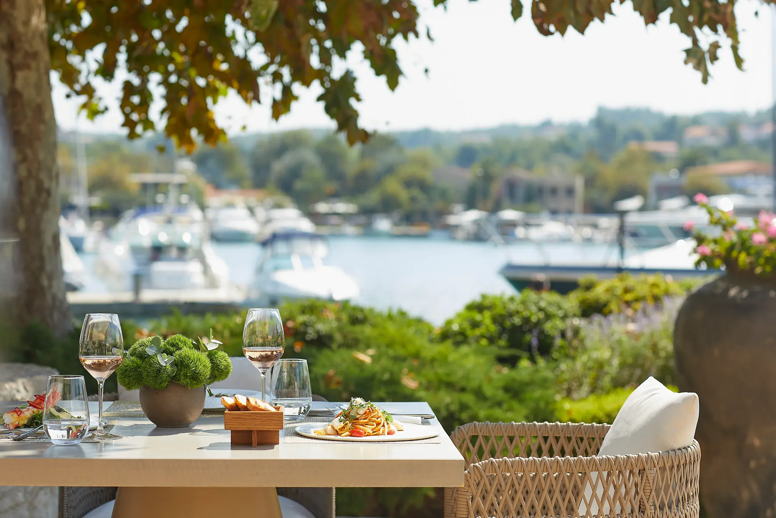 Waterfront outdoor dining table set with gourmet dishes and wine, overlooking a marina with boats on a sunny day.