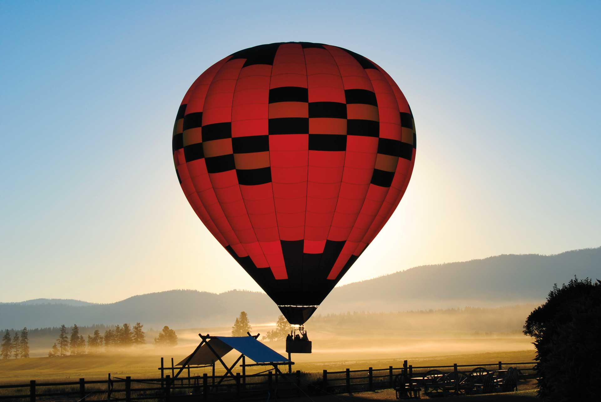 Red and black hot air balloon floating above a misty landscape at sunrise with rolling hills in the distance.