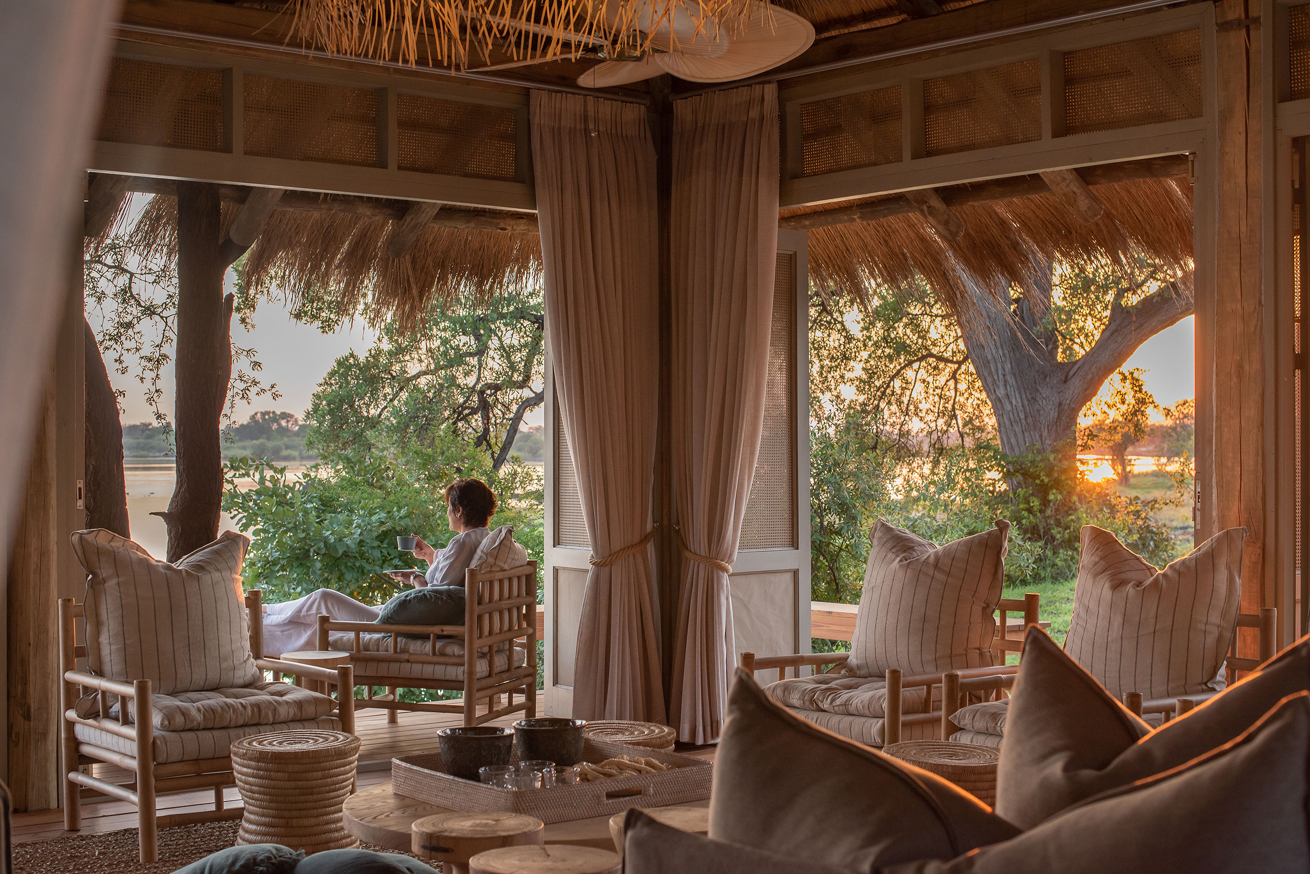 The plush neutral interior of a lounge area with a woman sat on the outdoor deck as the sun sets