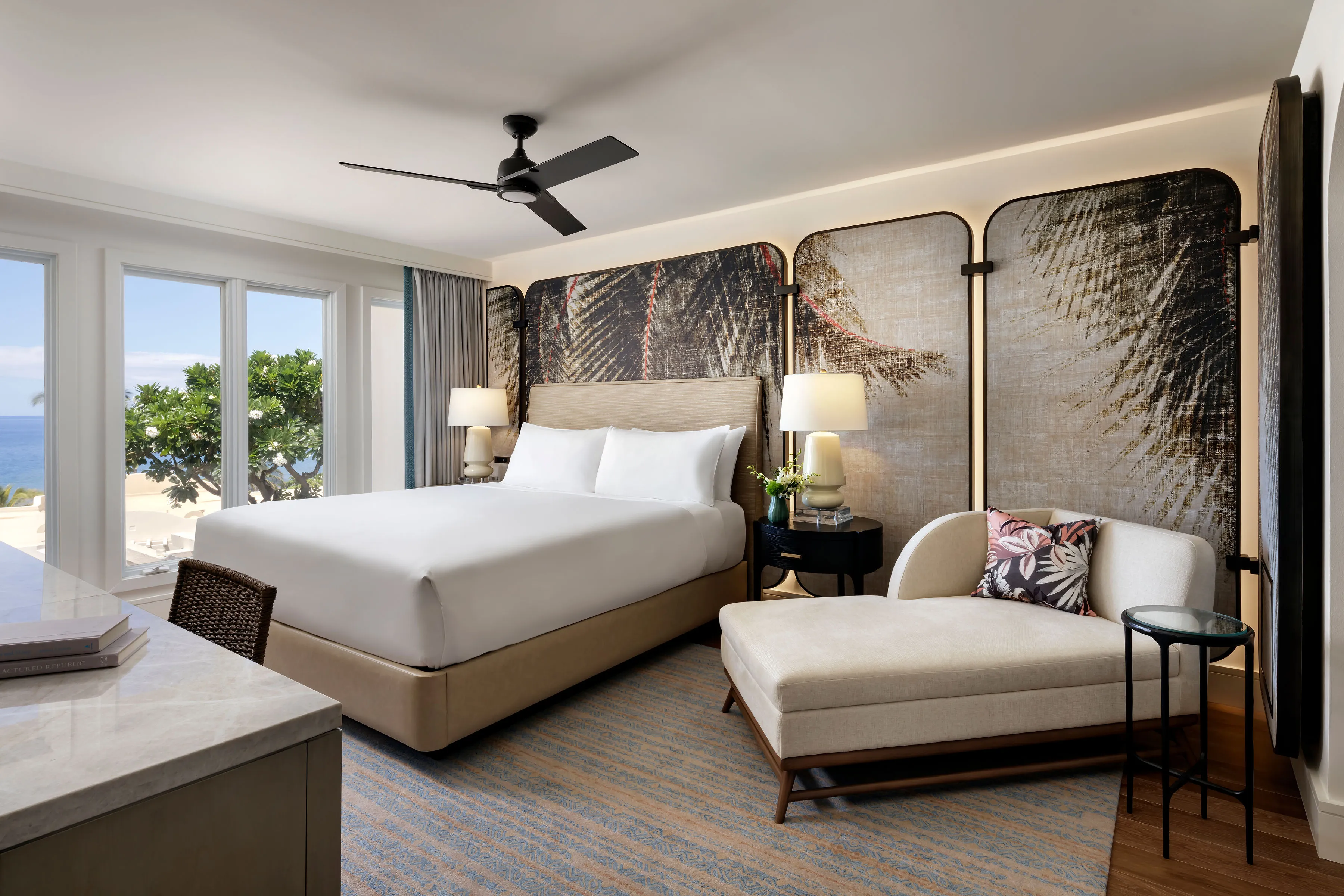 King size bedroom of a villa at Fairmont Kea Lani featuring a large bed in front of a woven palm tree mural, a pale chaise longue, and view of the ocean 