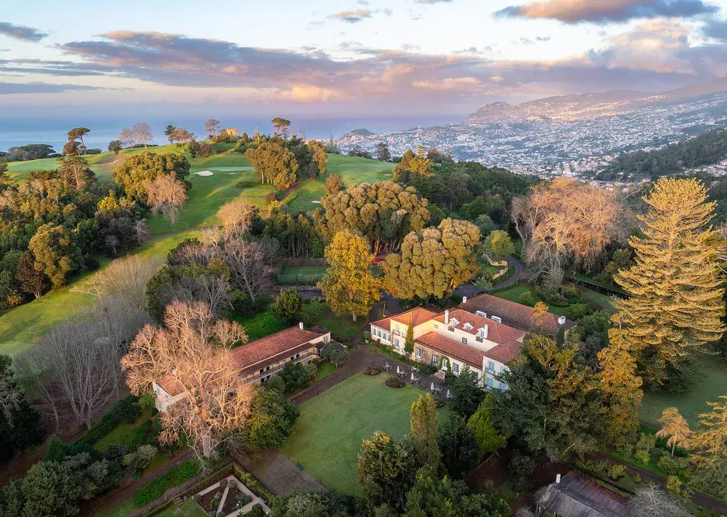 An aerial view of Casa Velha do Palheiro nestled among trees and a golf course overlooking Funchal city in the distance
