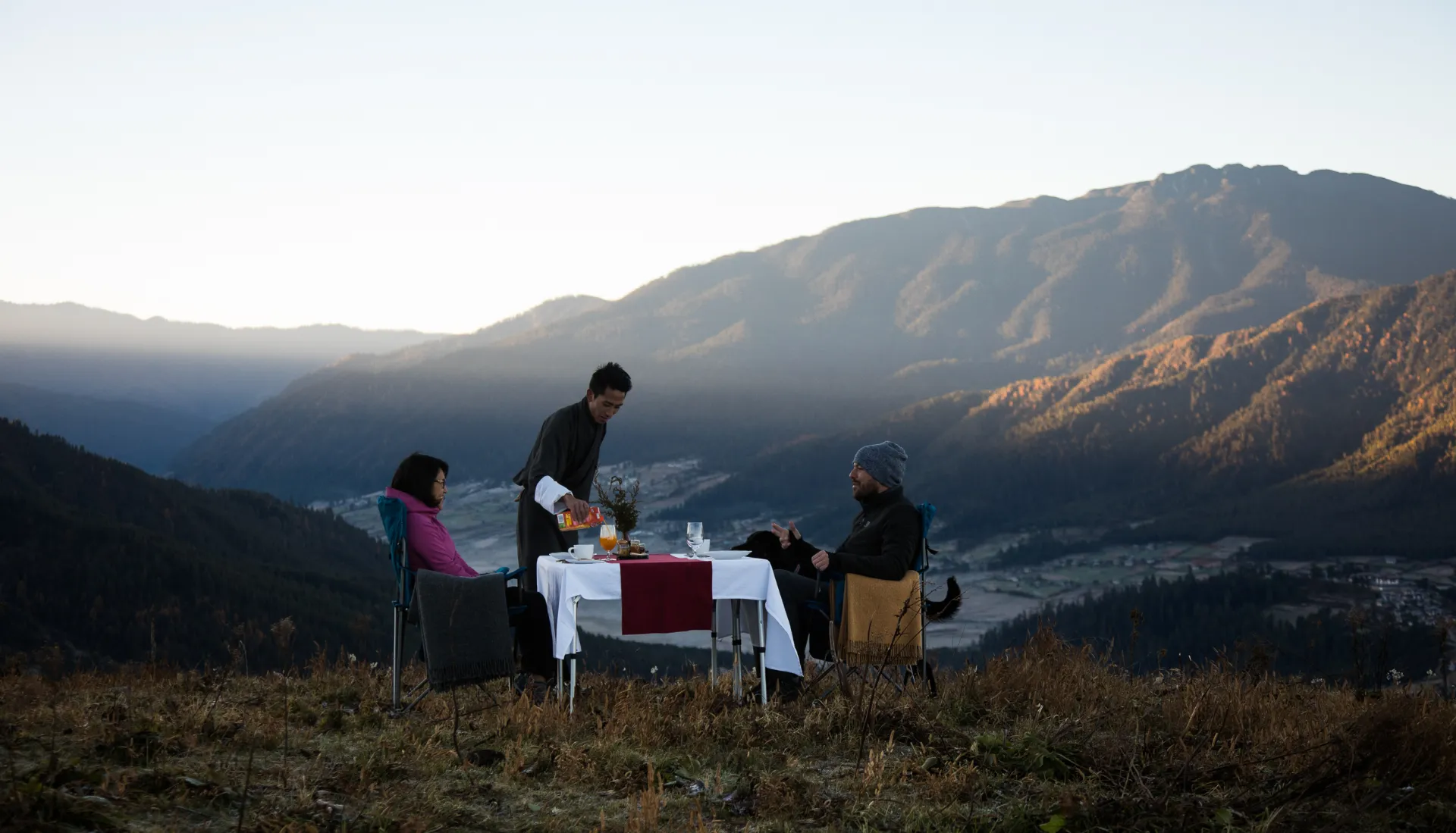 Outdoor dining setup on a mountain hillside with views of a sunlit valley and peaks.