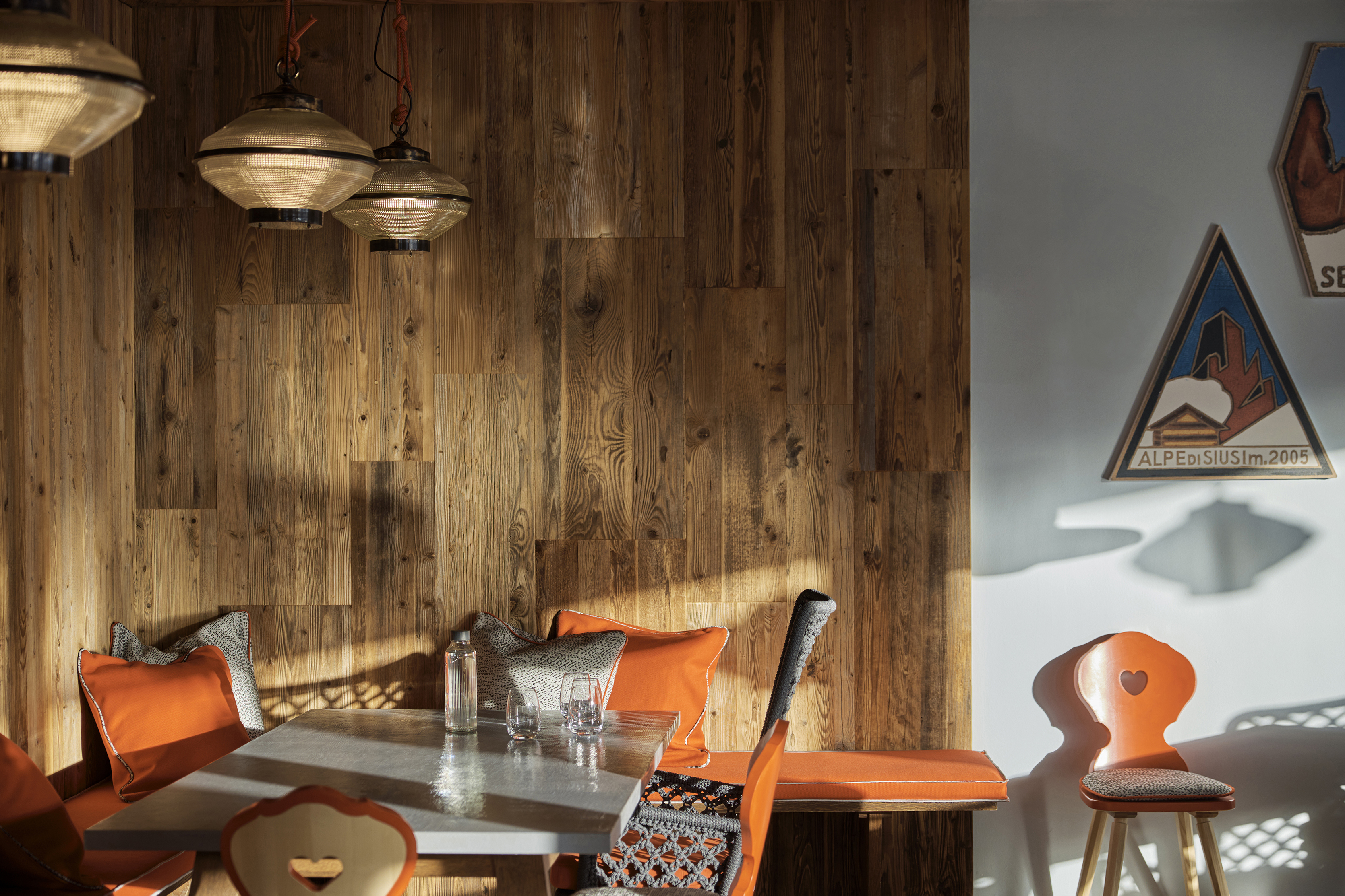 A cafe-style corner with orange chairs, patterned cushions, and wooden walls illuminated by natural light and hanging lamps.