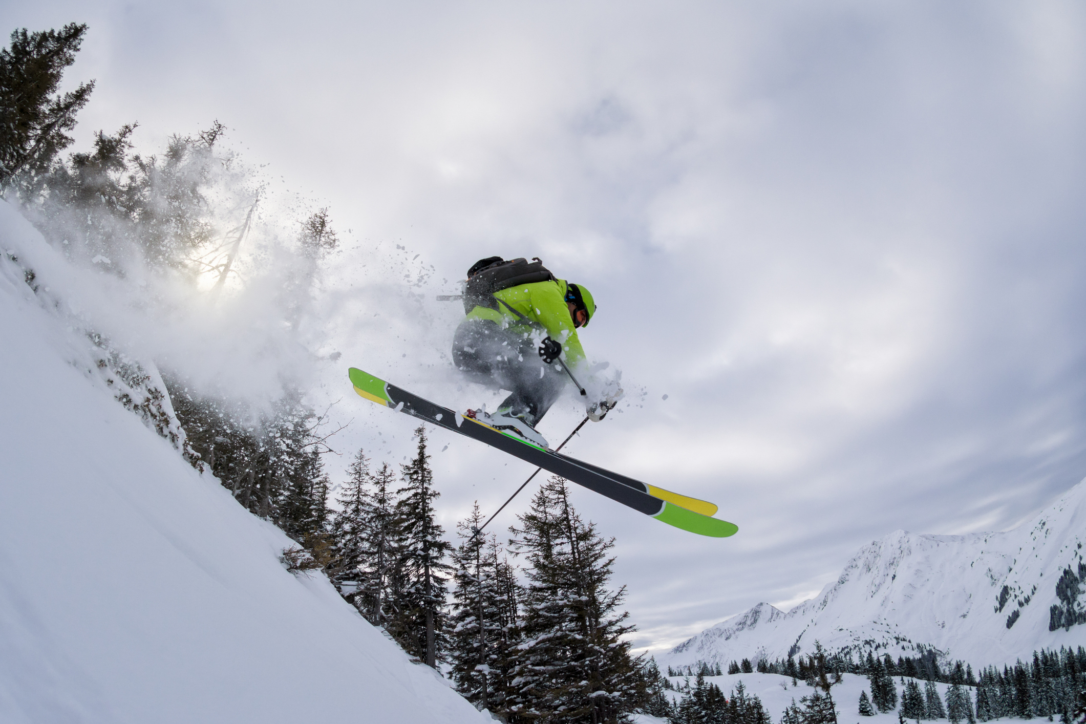 A skier in mid-jump against a snowy mountain backdrop with evergreen trees and a cloudy sky.