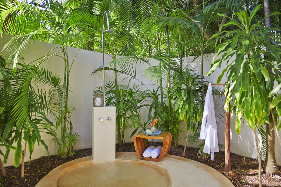 An outdoor shower with white towel and wooden furniture surrounded by greenery and a white wall for privacy