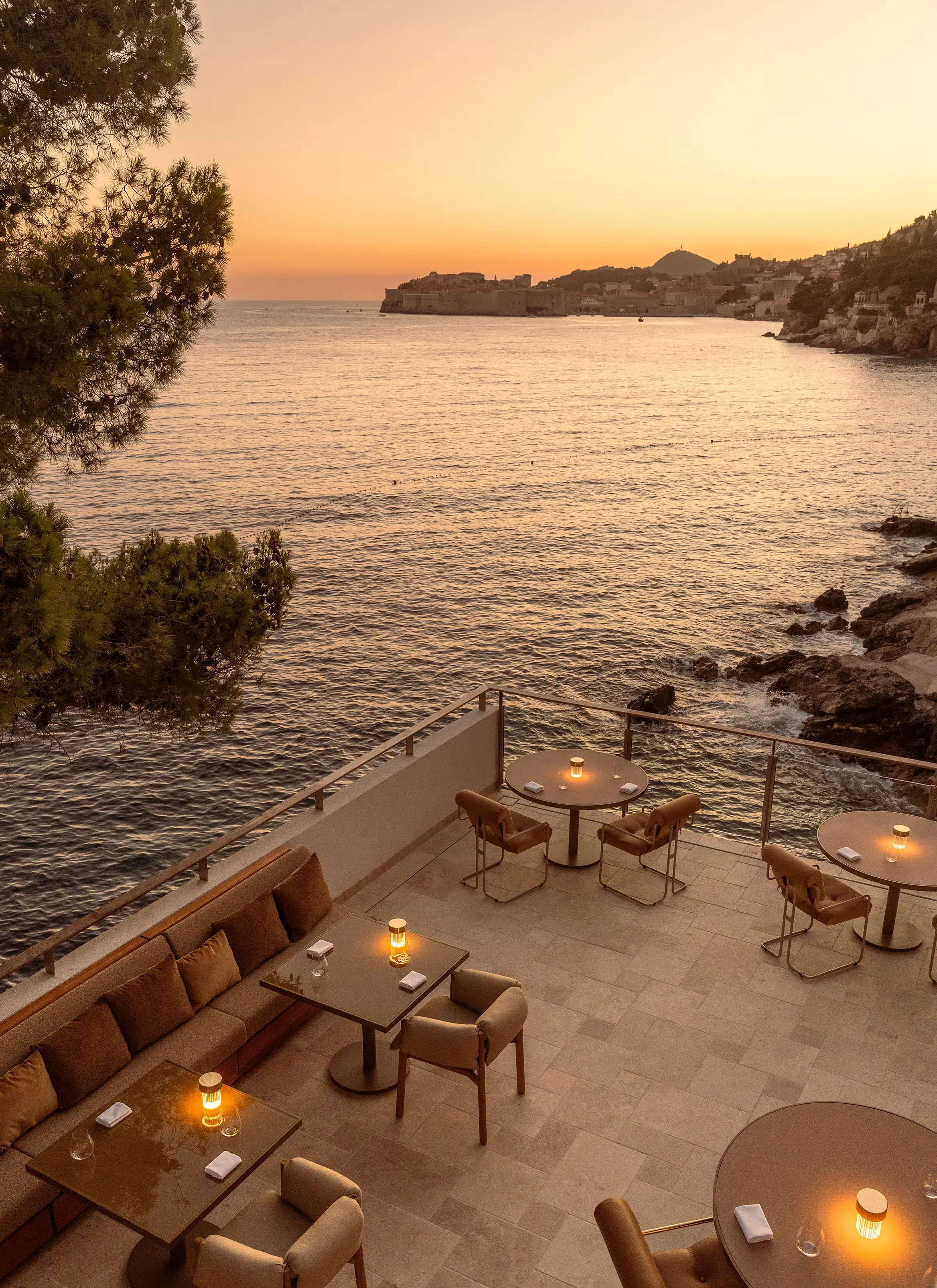 Dining terrace of Pjerin Restaurant at Villa Dubrovnik in Croatia during sunset, featuring tables set for dinner with lamps beside the water and a view of the Old Town behind
