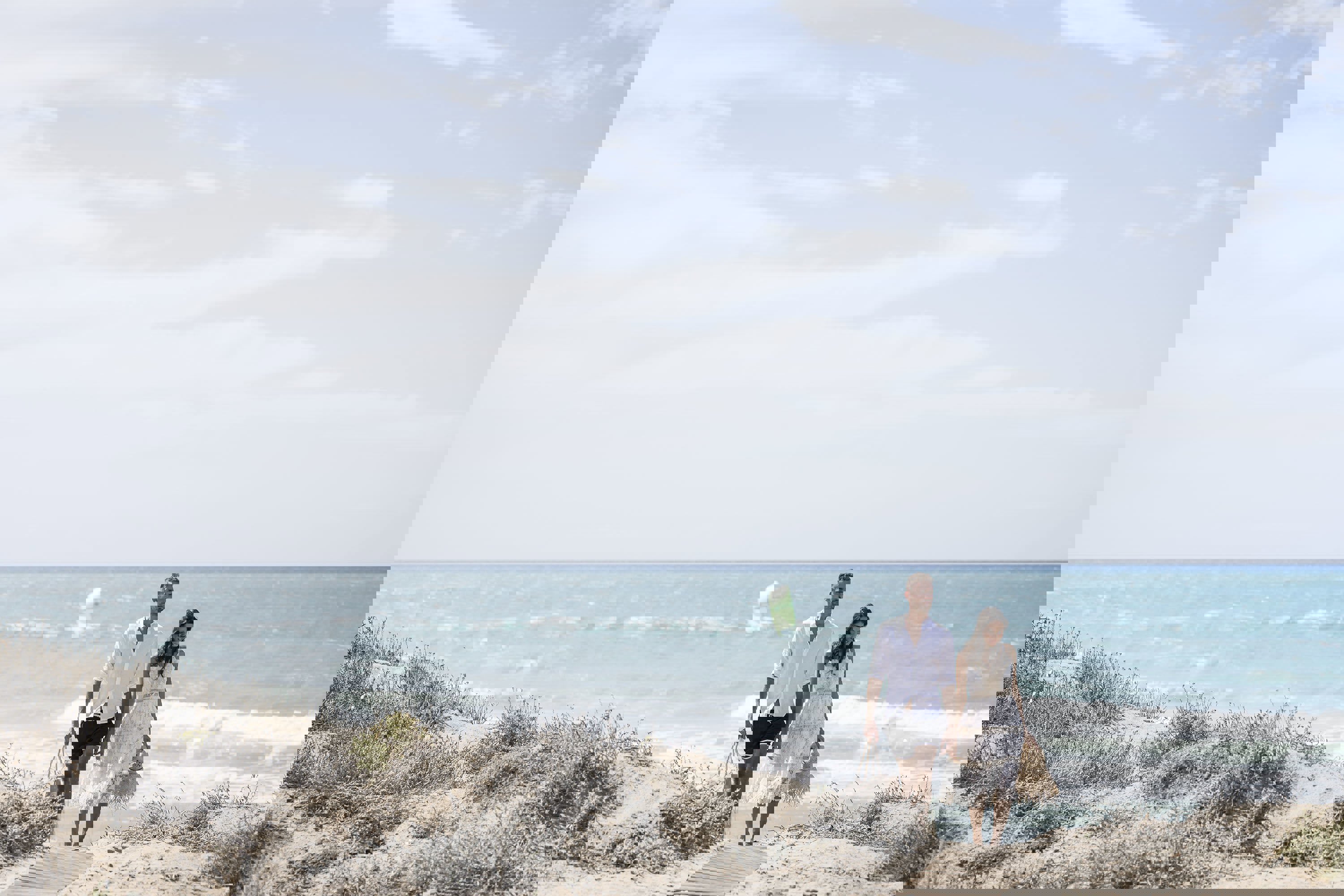 Couple walking hand in hand up a soft sand beach with a kite surfer on the sea in the background