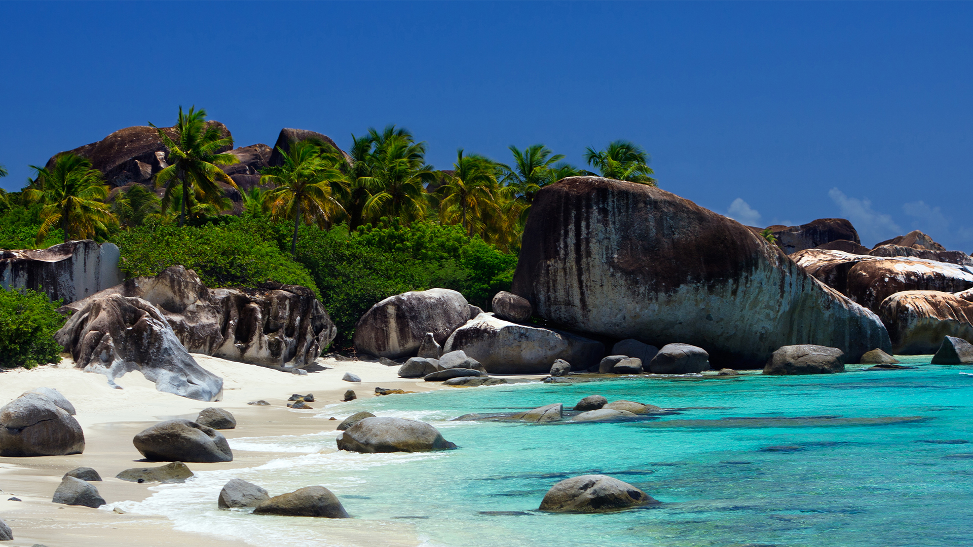 A tropical beach with clear turquoise waters, white sand, and large granite boulders under a blue sky.