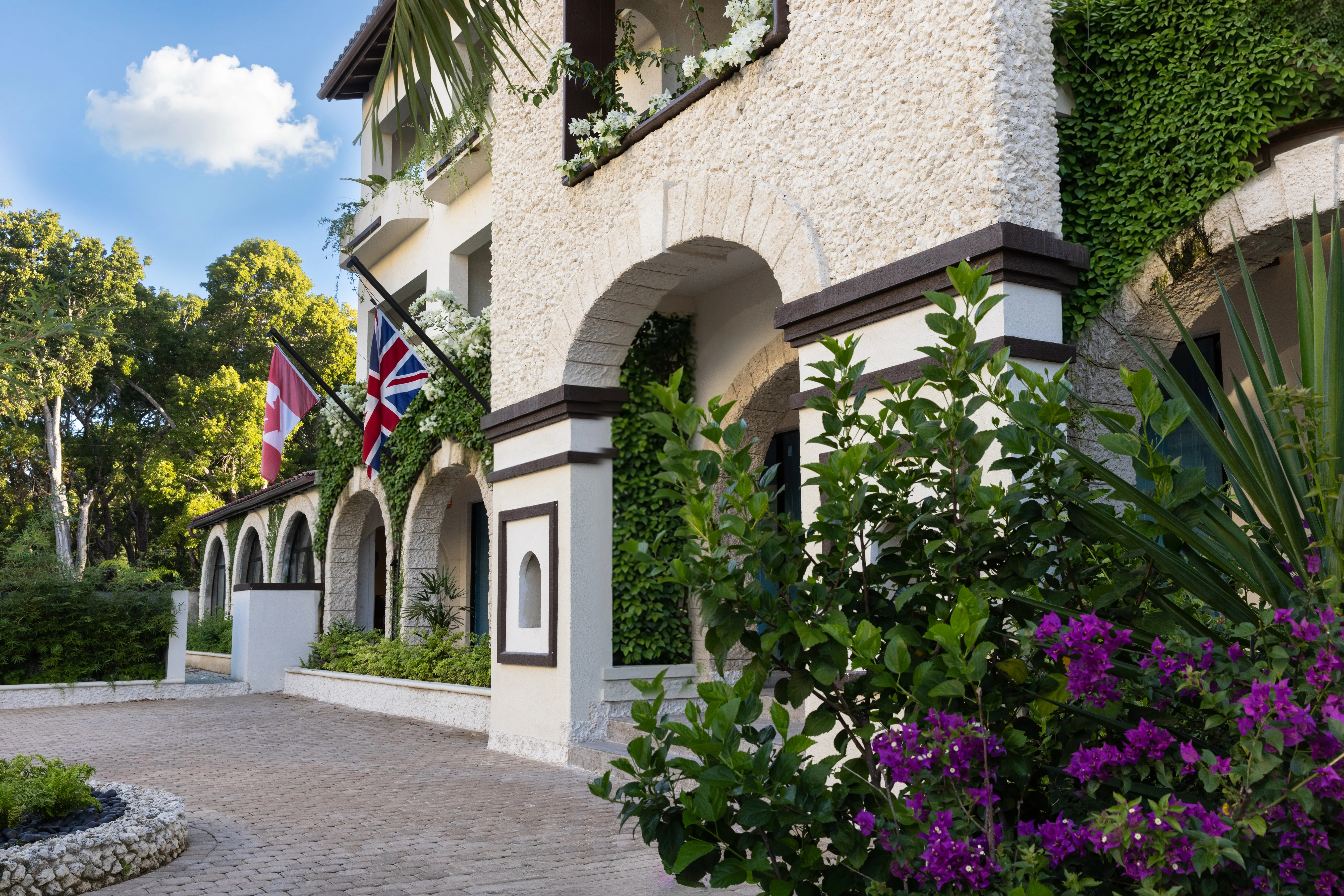Mediterranean-style building with arched walkways, lush landscaping, and Canadian and UK flags displayed at the entrance.