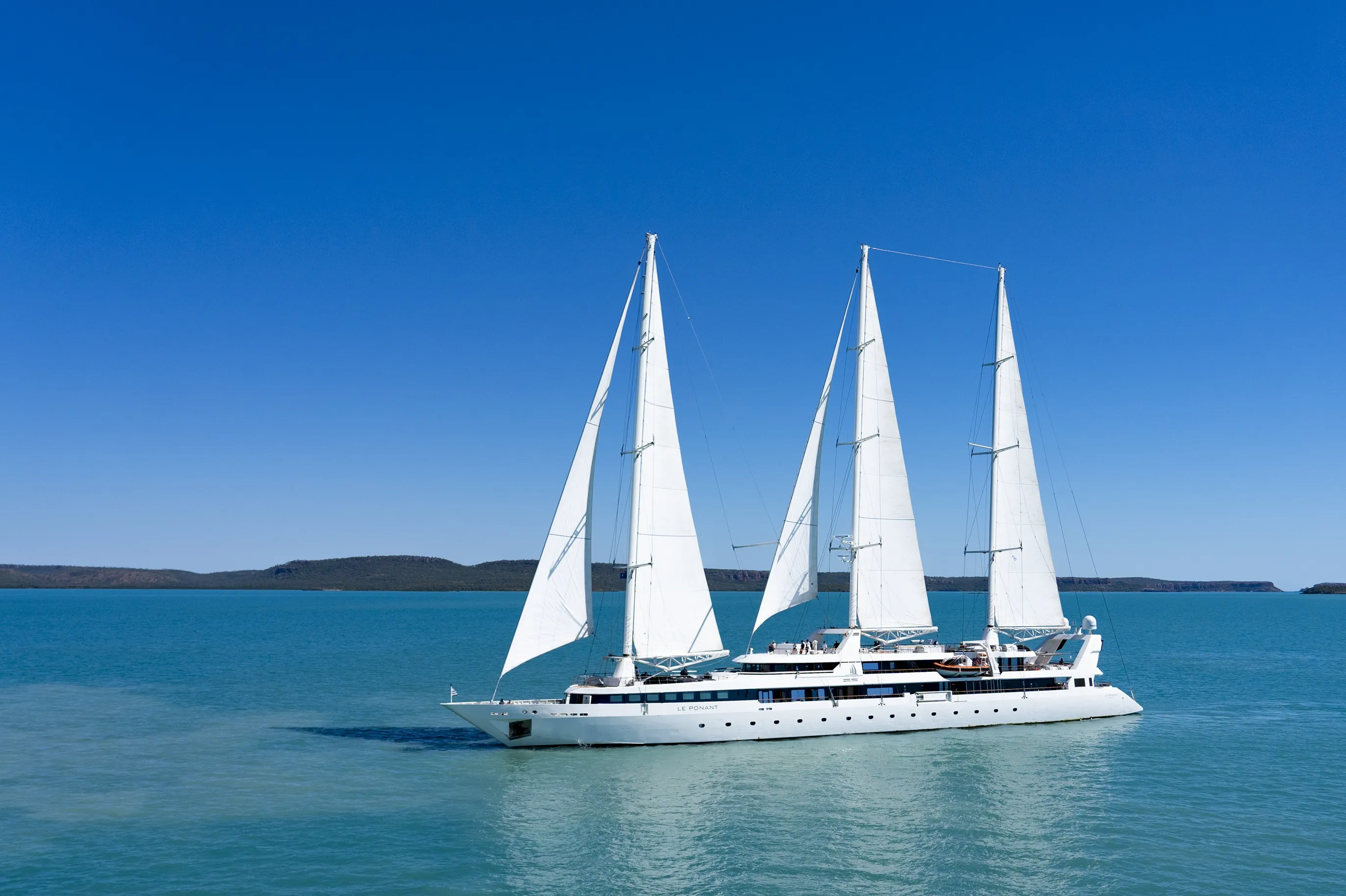 Three-masted white PONANT sailing yacht gliding across calm blue waters with distant land on the horizon.