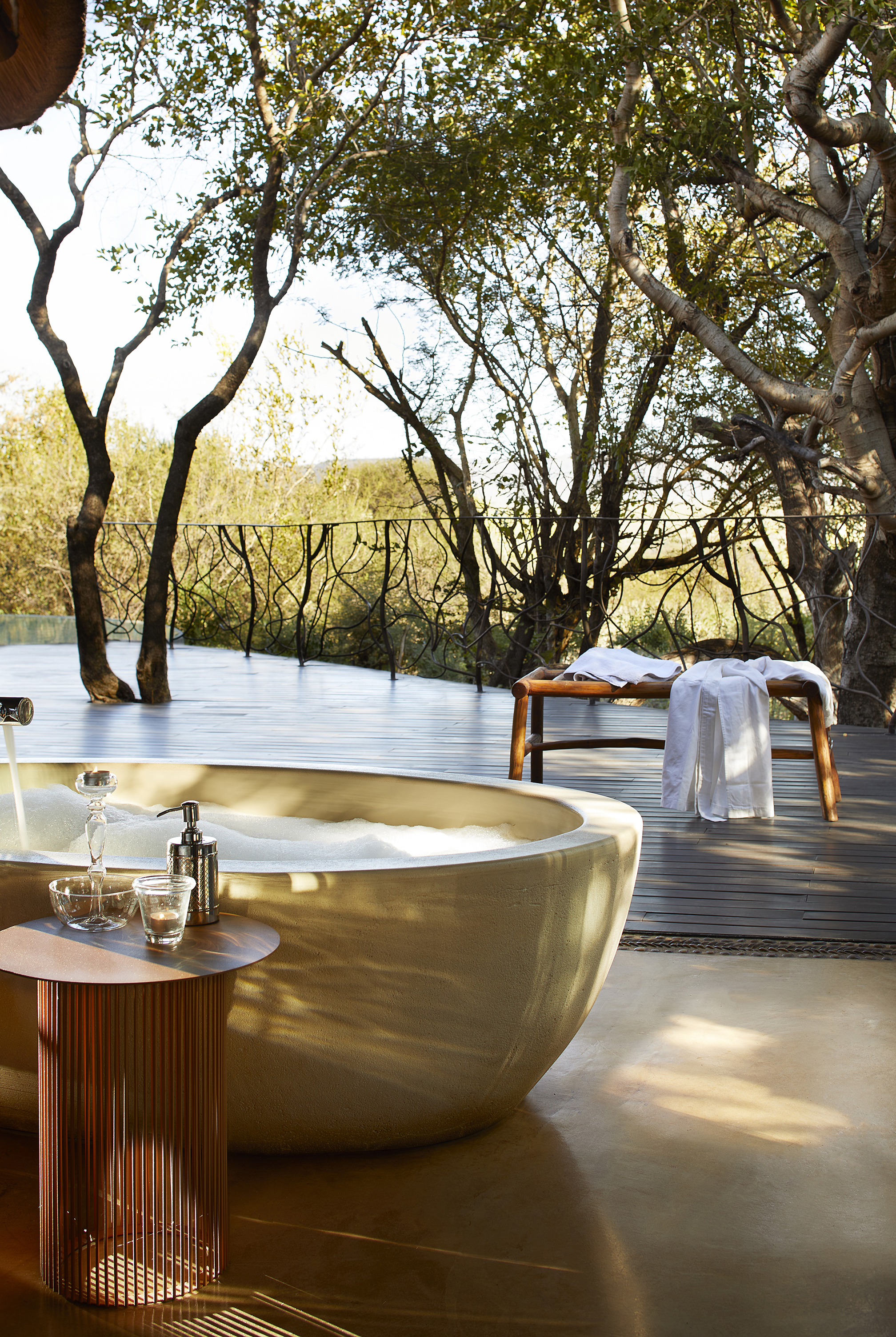 An open air bathroom terrace surrounded by trees and featuring a deep stone stand-alone bathtub, a bench with towels and a table with soap and a candle
