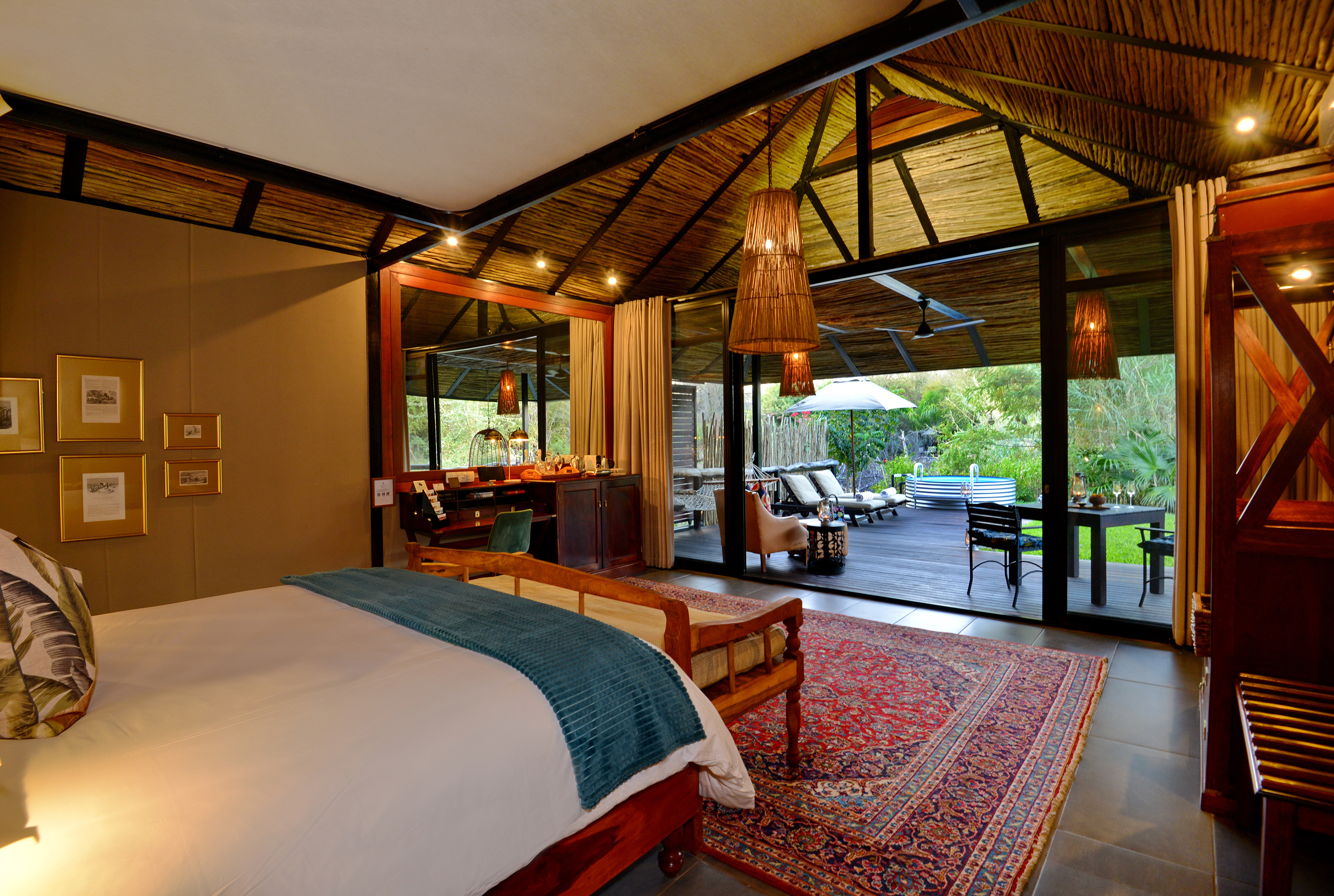 A luxurious yet rustic bedroom at Mukwa River Lodge featuring a large bed with white linens and teal throw, wooden furniture, and an open view to a spacious outdoor patio with seating and greenery.