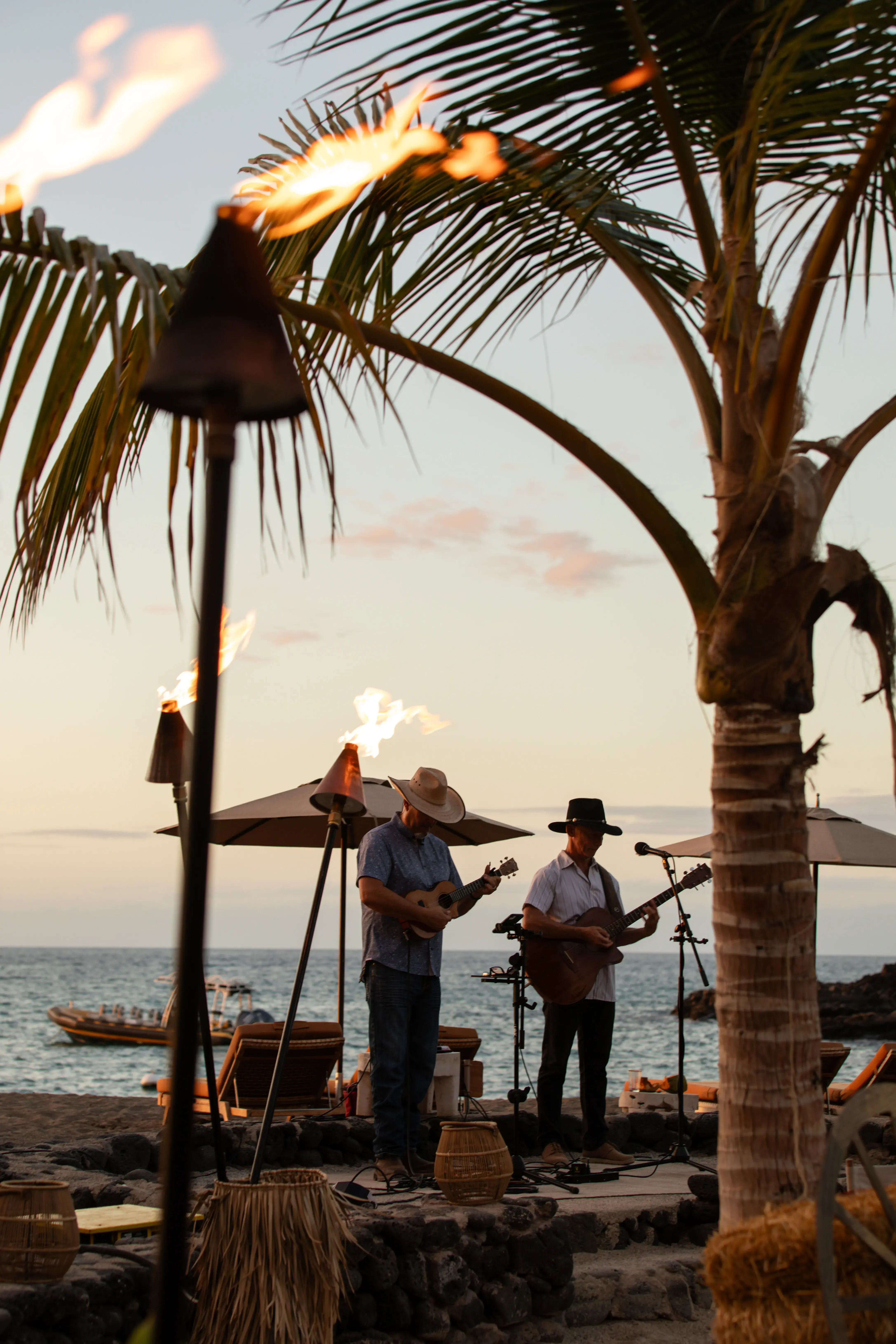 Two musicians performing on an oceanside stage surrounded by palms, torches and sun loungers