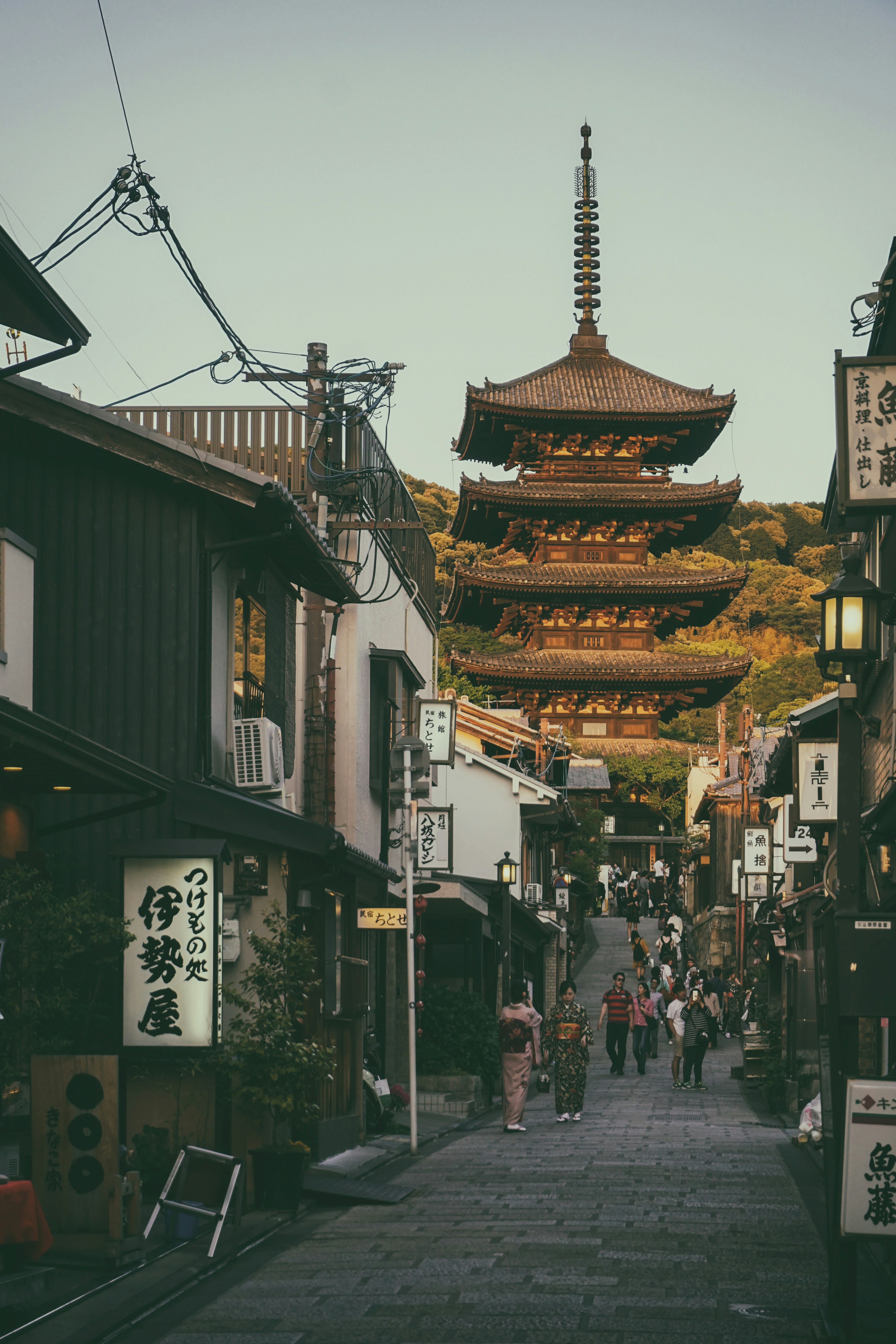 A cobbled street in Kyoto lined with old buildings leading to a pagoda with people walking including two geishas