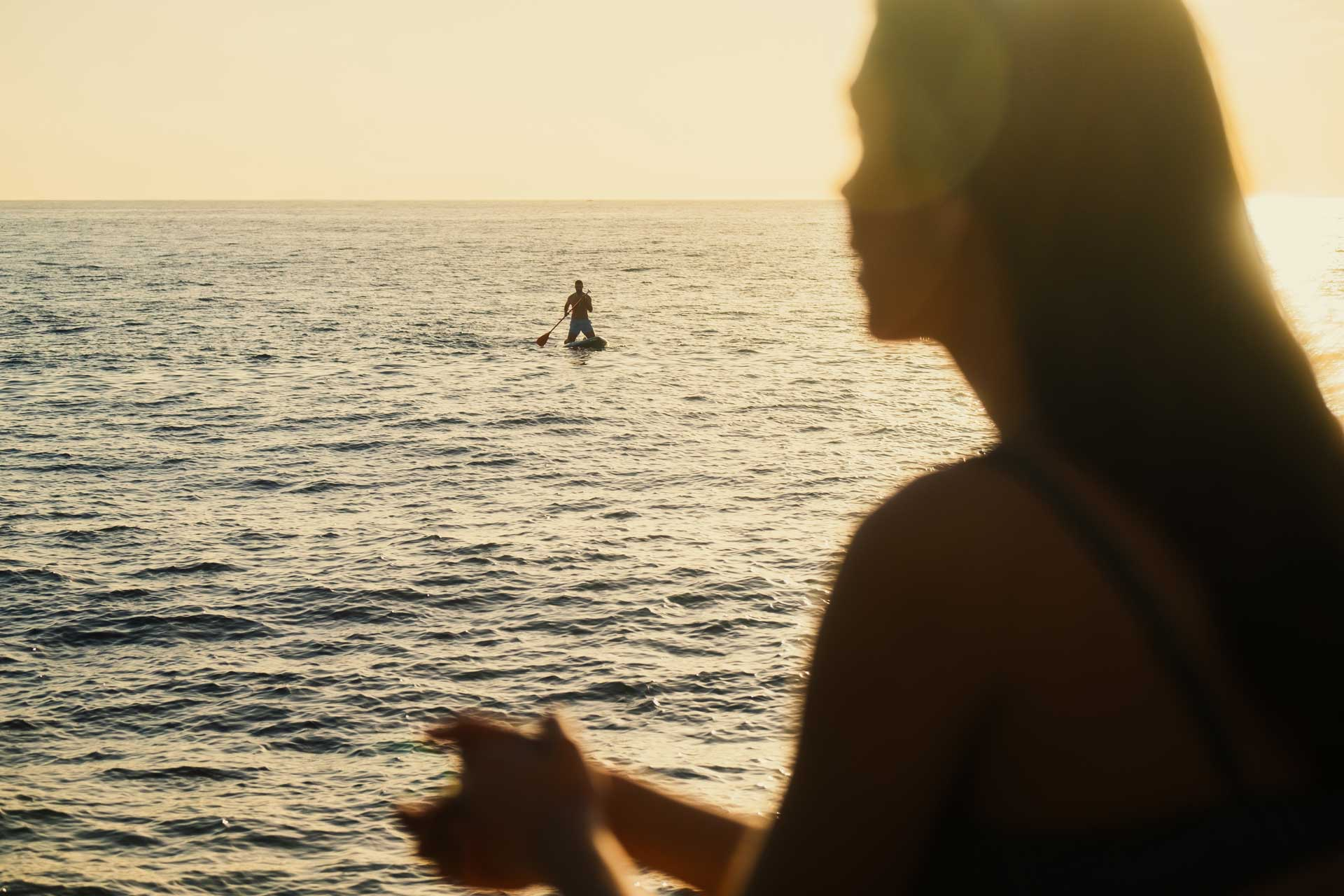 Silhouetted person in the foreground watching someone paddleboarding on calm sea waters at golden hour.