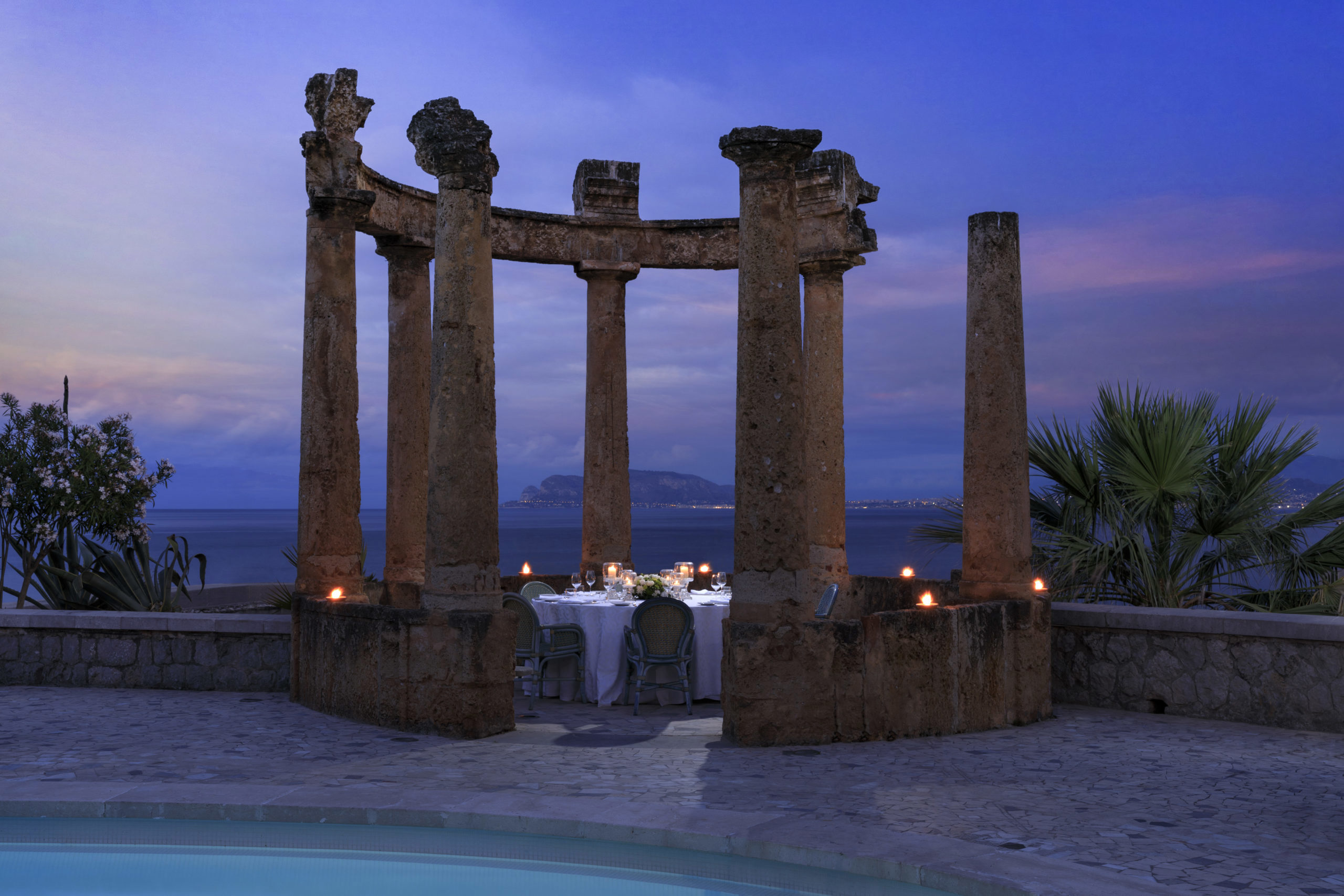 An evening view of a romantic outdoor dining setup with lit candles, surrounded by ruined columns overlooking a calm sea and distant lights on the horizon.